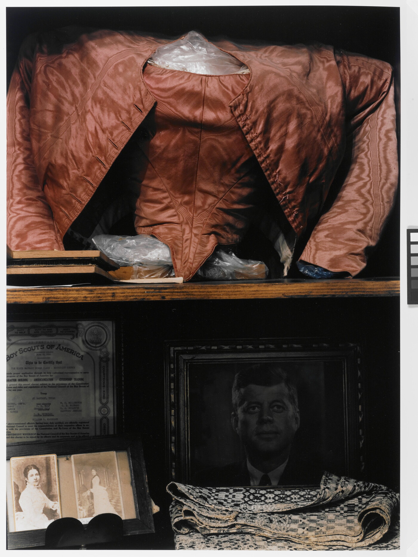 A color photograph of a shelf displaying a red shirt and jacket, photographs of two women, a portrait of John F. Kennedy, some folded fabric, and a Boy Scouts of American certificate.