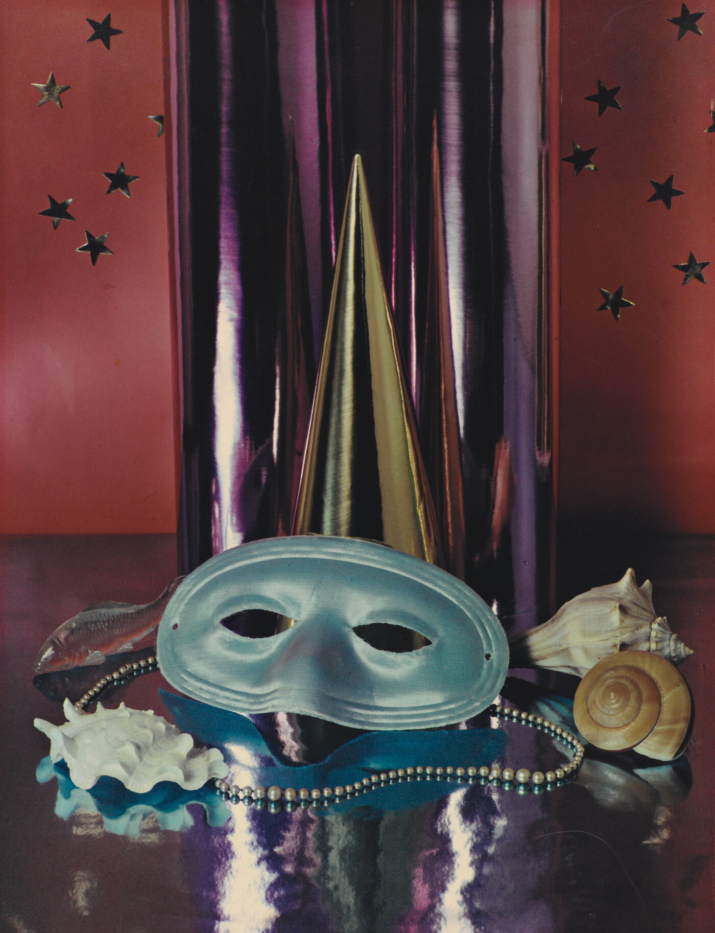 A color photograph of a simple party mask, Mardi Gras beads, and seashells in front of a reflective silver cylinder.