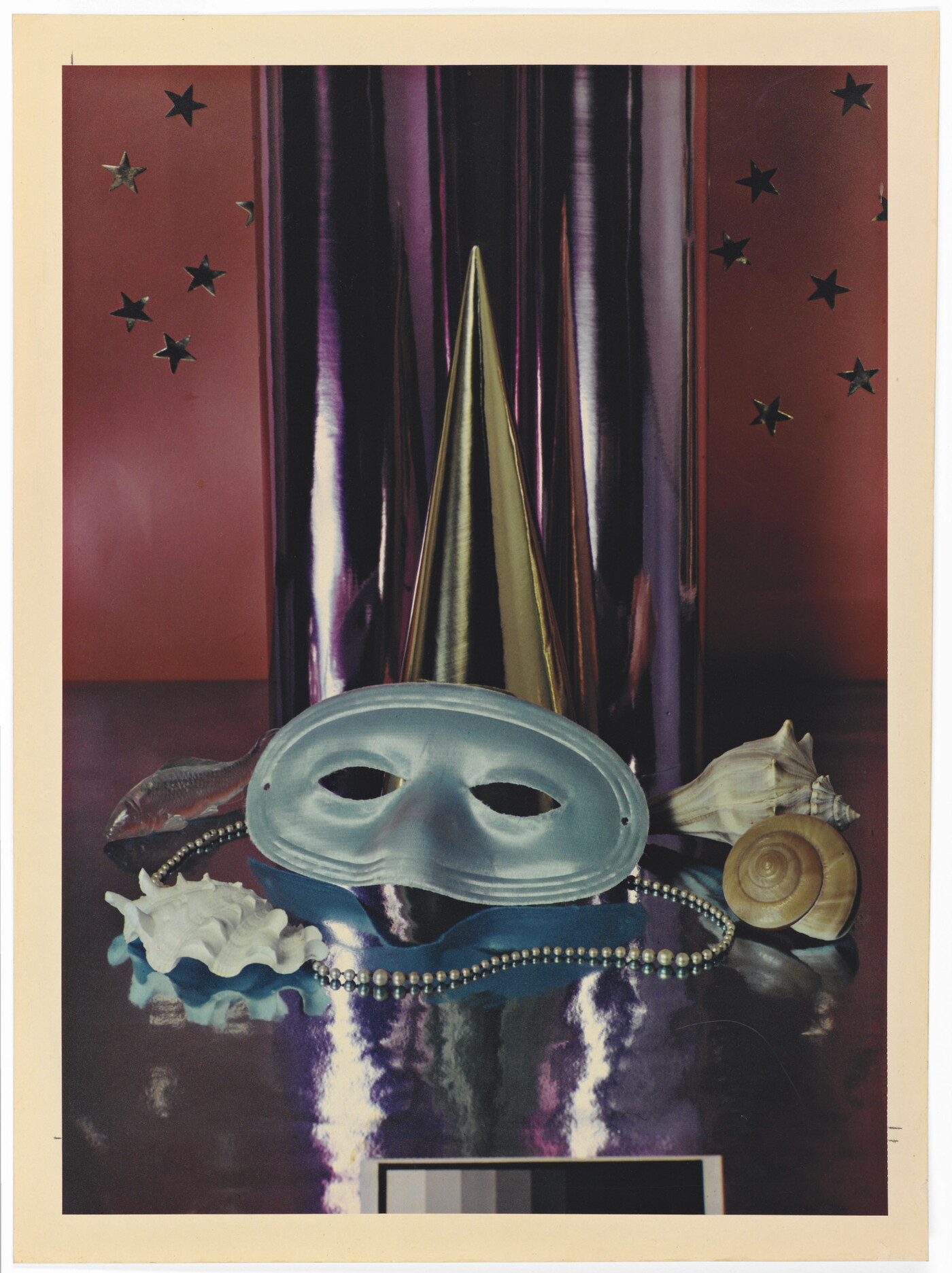 A color photograph of a simple party mask, Mardi Gras beads, and seashells in front of a reflective silver cylinder.