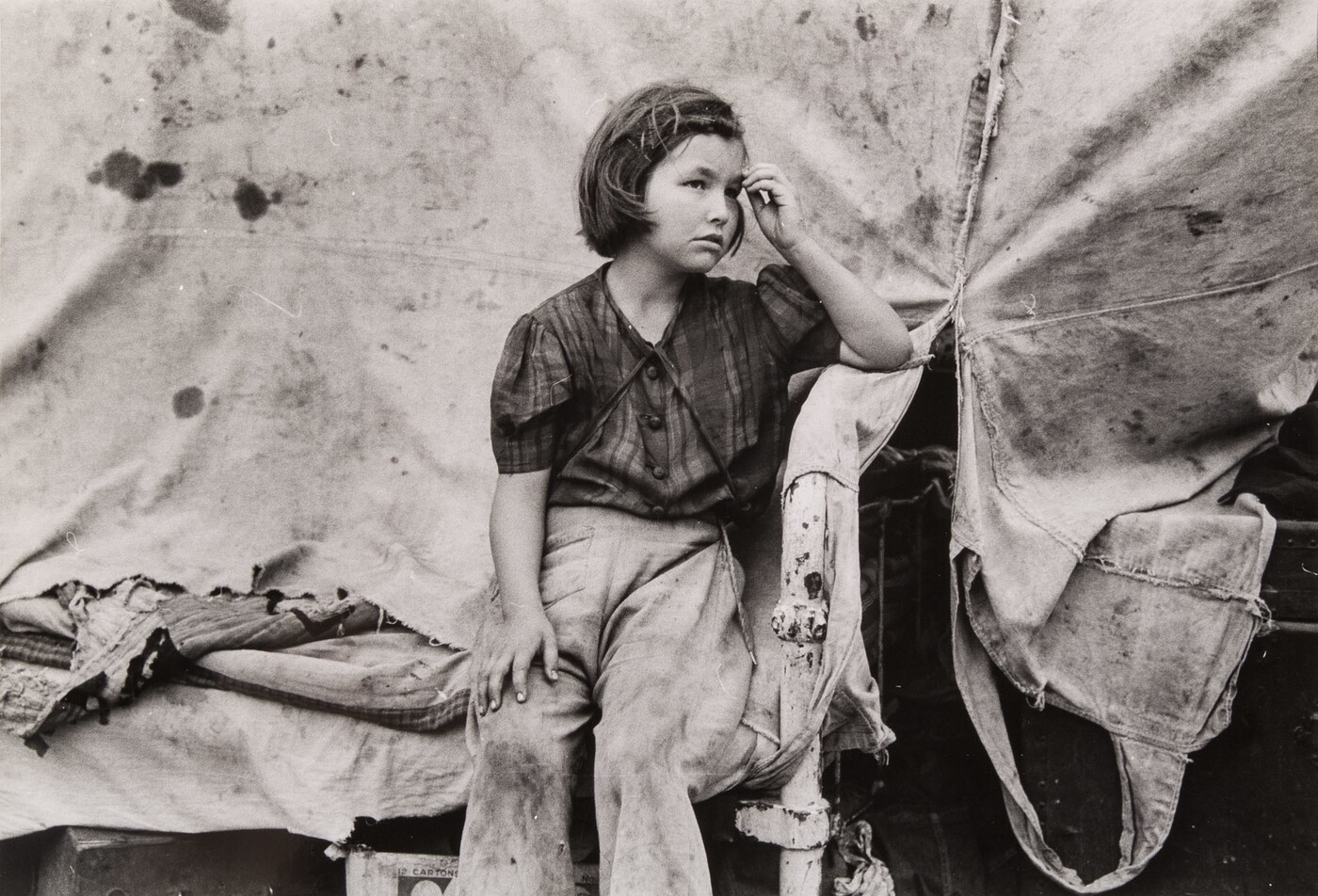 A black-and-white photograph of a young White girl wearing dirty clothes, one hand held up to her forehead, sitting in front of a canvas tent.