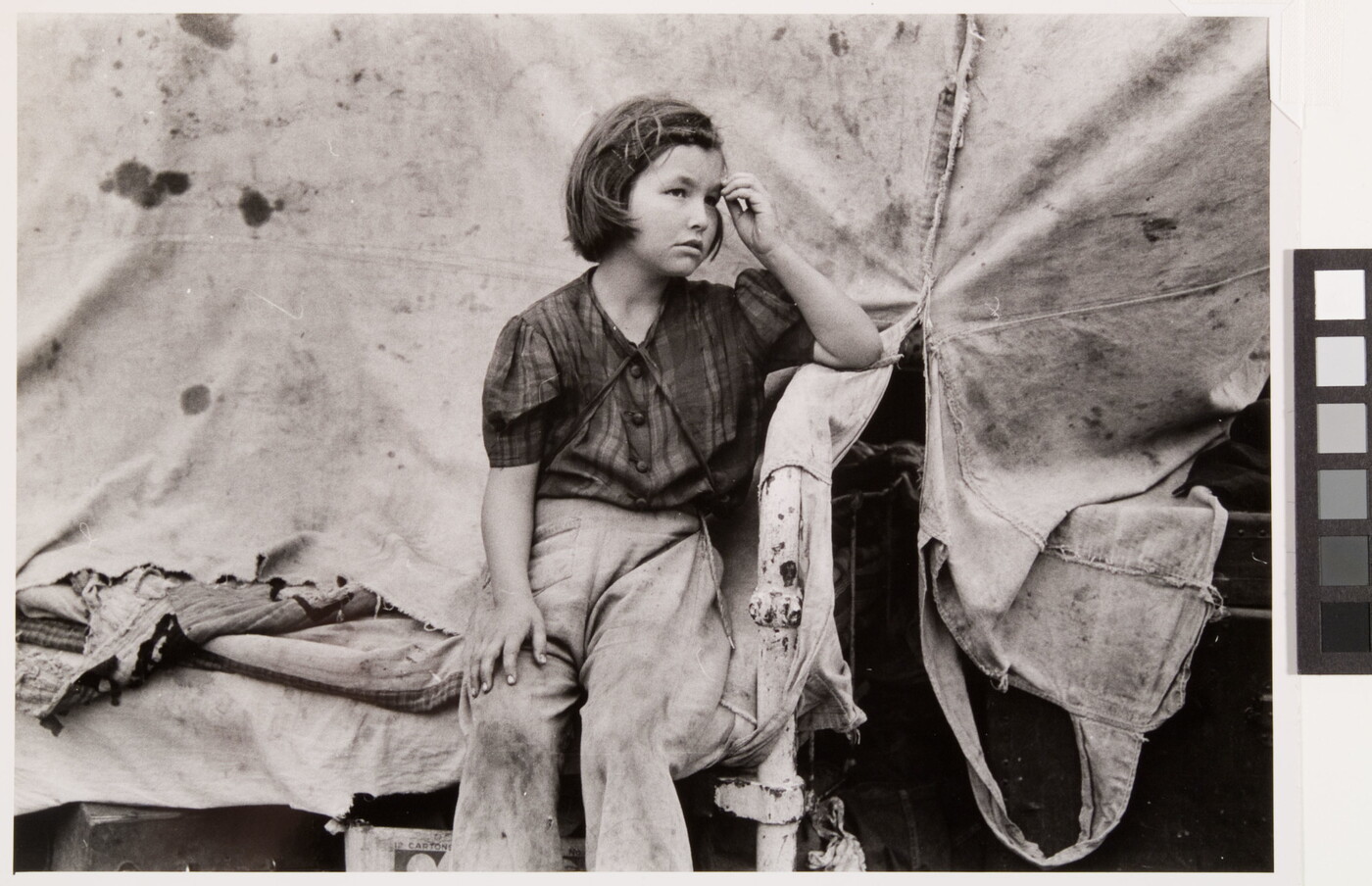 A black-and-white photograph of a young White girl wearing dirty clothes, one hand held up to her forehead, sitting in front of a canvas tent.