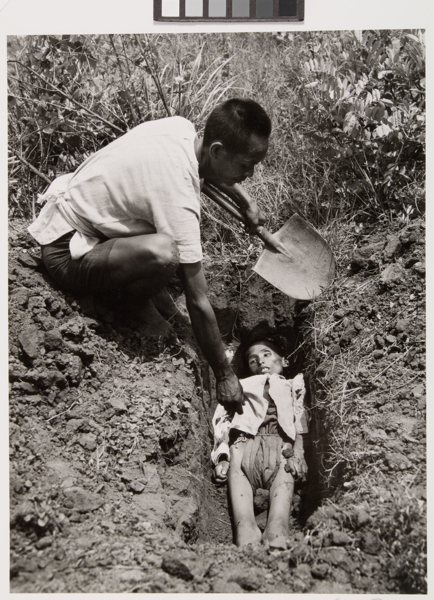 A black-and-white photograph of an Asian man holding a shovel crouched down next to a freshly dug grave containing a emaciated body.