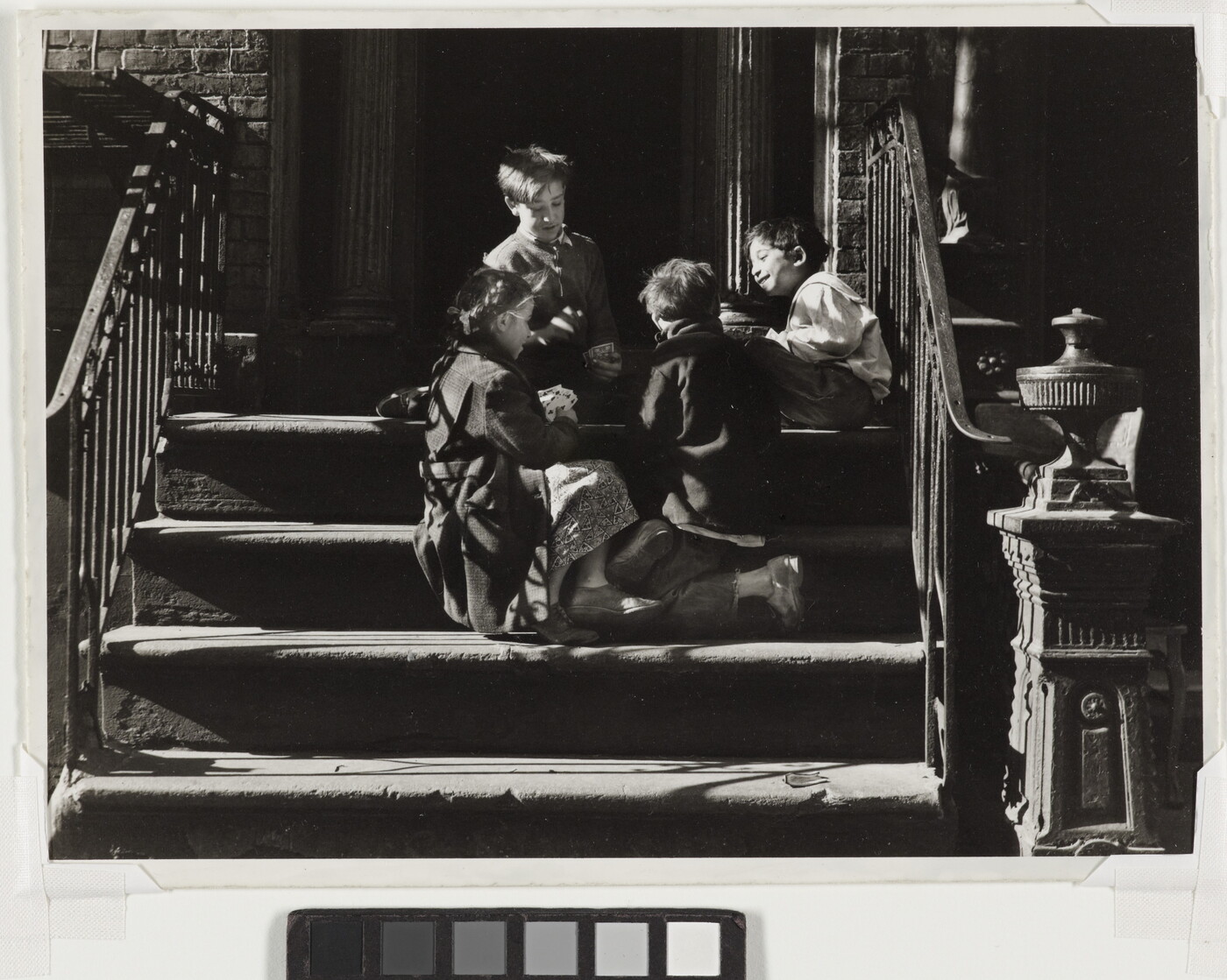 A black-and-white photograph of four children playing together on concrete steps with iron railings that cast striped shadows across them.