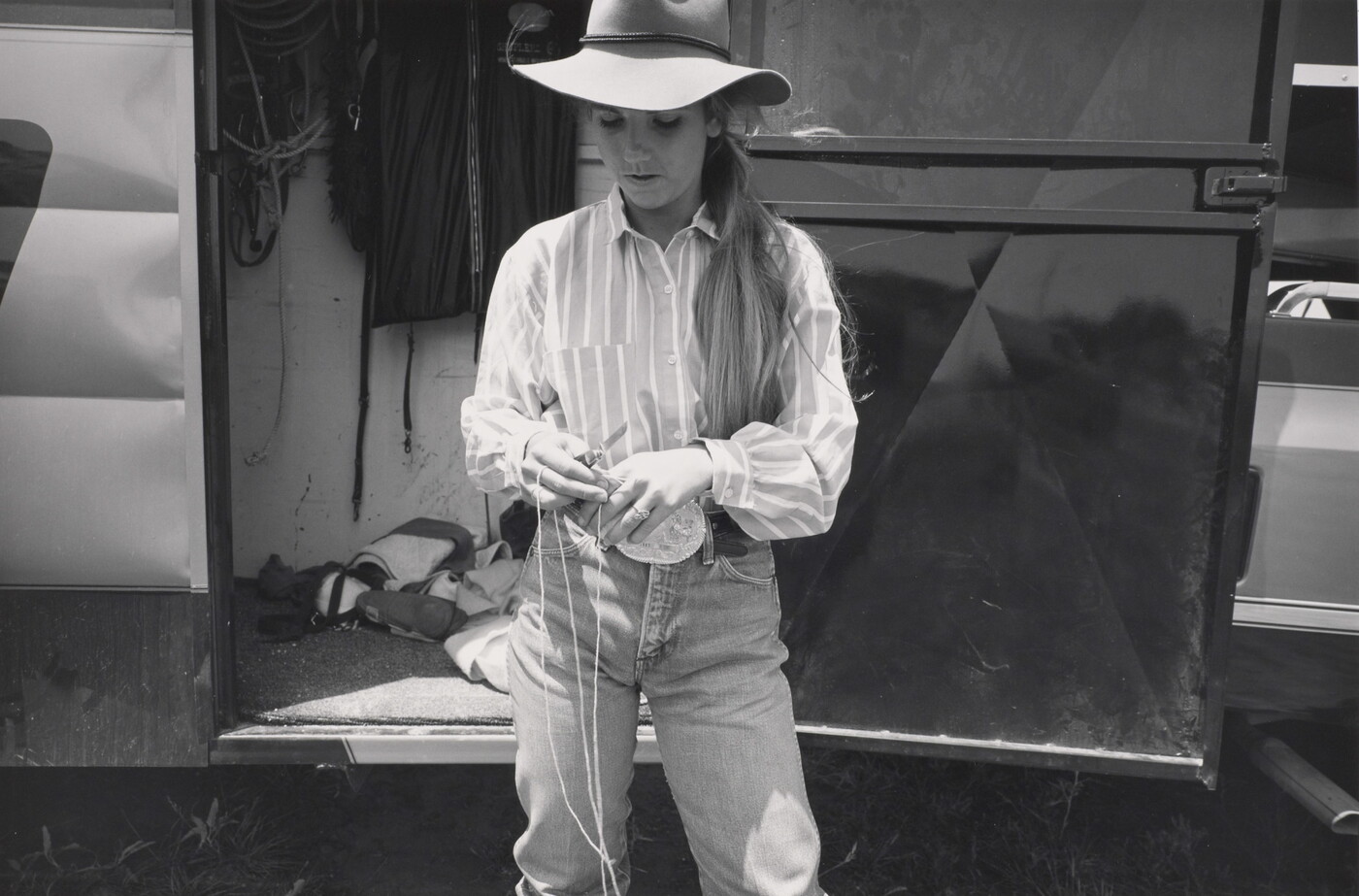 A black-and-white photograph of a young White woman with long hair wearing western clothing cutting rope with a knife.