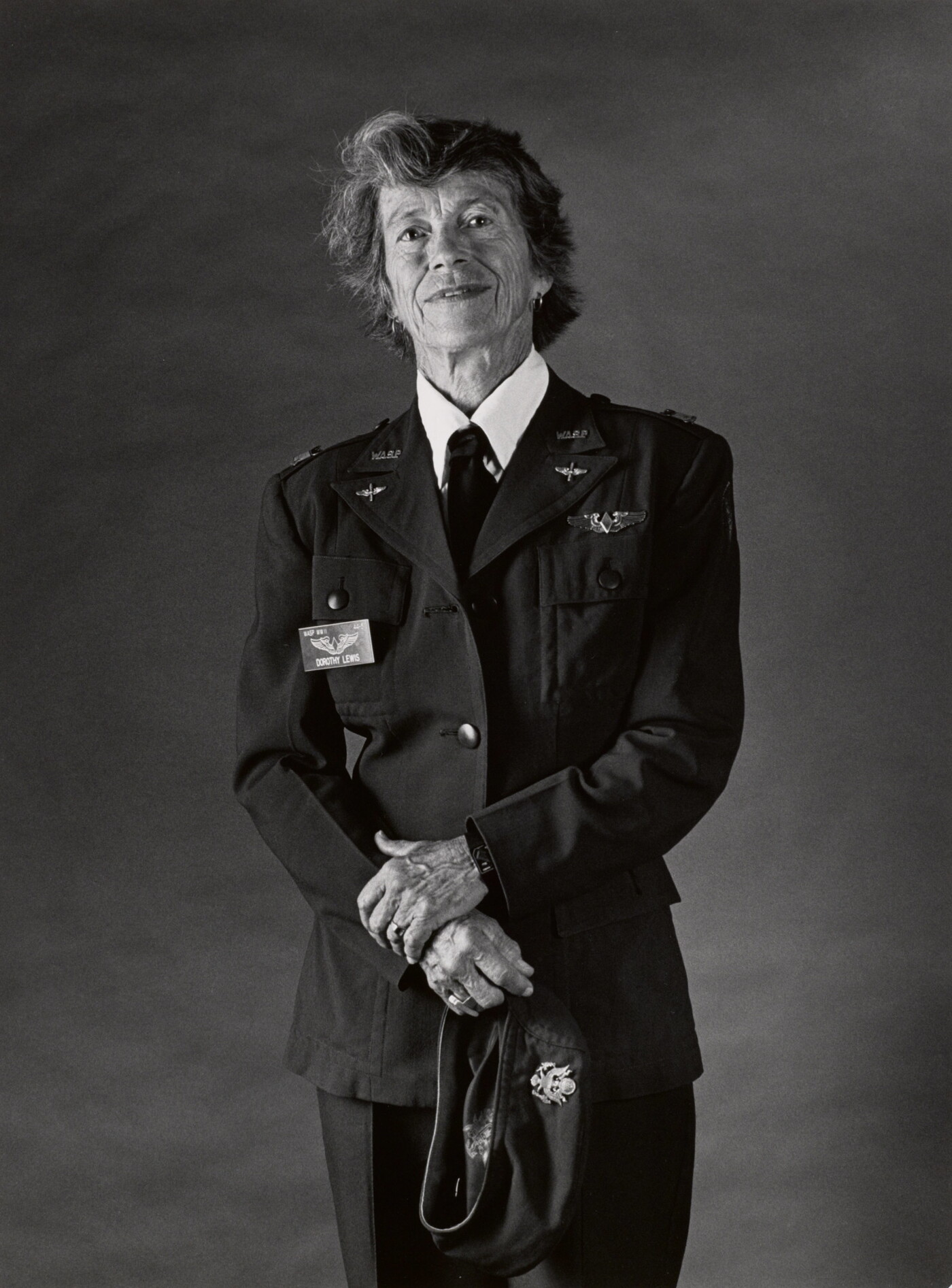 A black-and-white portrait photograph of an older White woman with short hair wearing a WASP military uniform and her hands in front of her holding a hat.