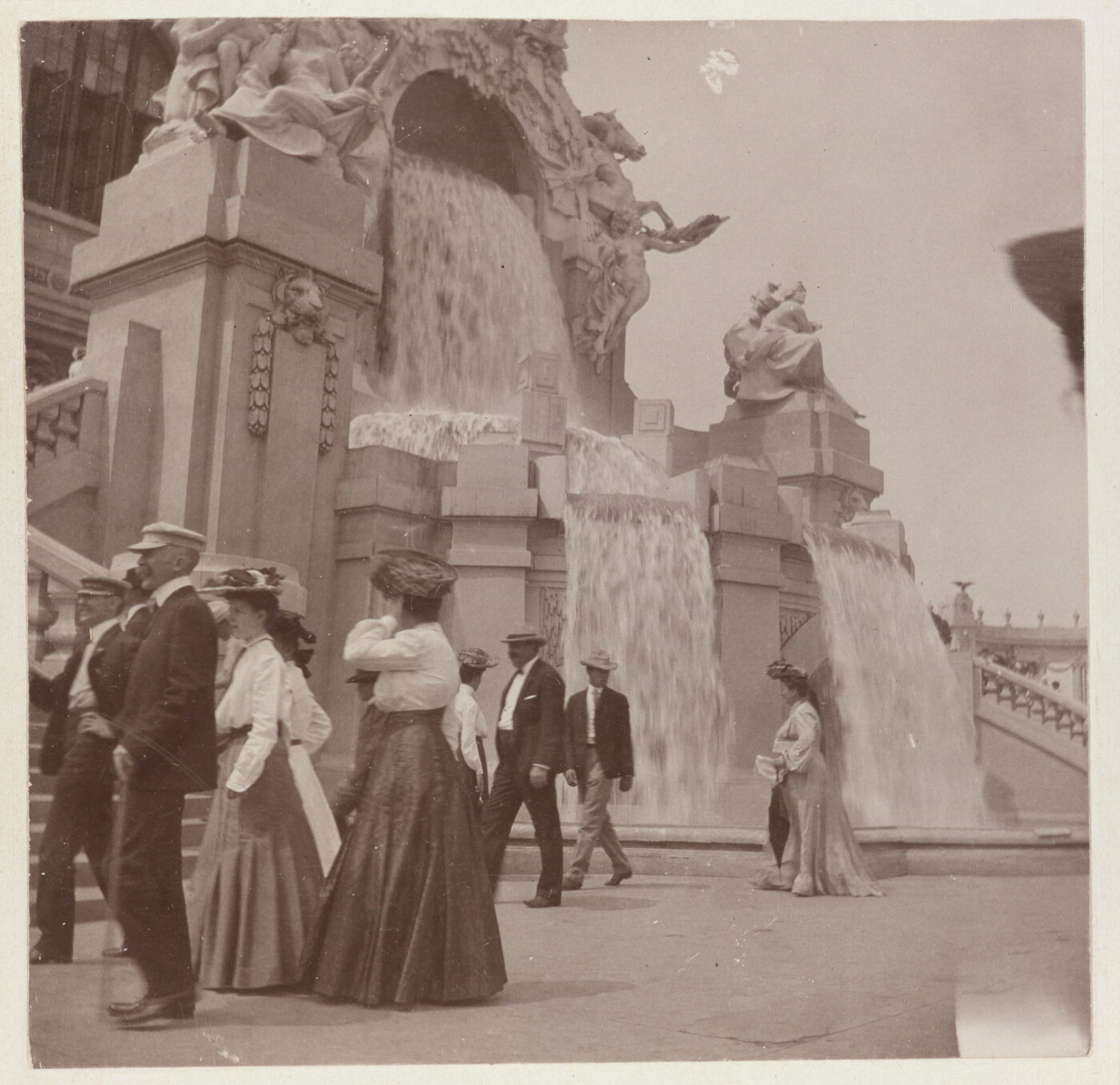 A sepia-toned photograph of women in long skirts and men in hats and jackets strolling by an elaborately sculpted facade with waterfalls.
