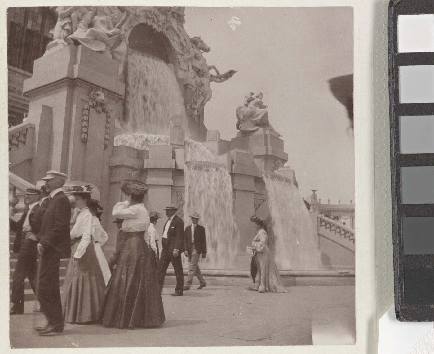 A sepia-toned photograph of women in long skirts and men in hats and jackets strolling by an elaborately sculpted facade with waterfalls.