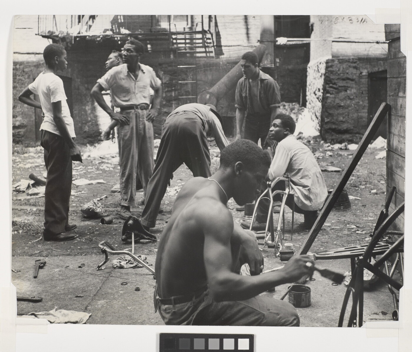 A black-and-white photograph of group of Black men talking and working in a rundown space littered with debris.
