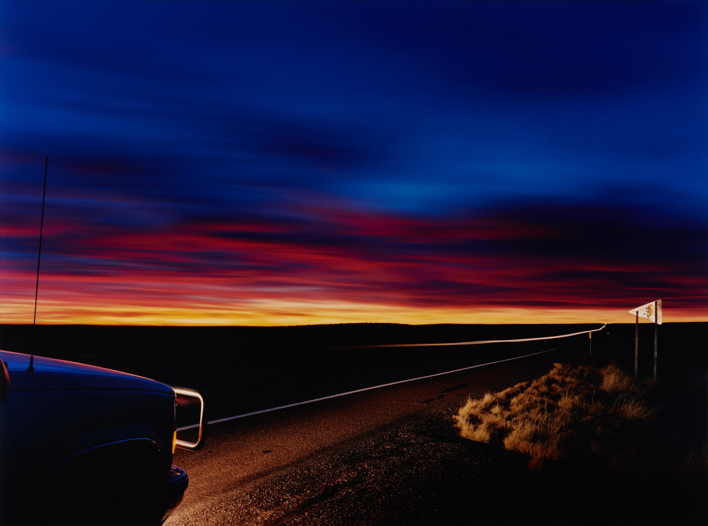 A color photograph of a deep blue sky over a line of orange and yellow sunset in a flat landscape; the lights from the front of a truck in the left corner illuminate part of the road.