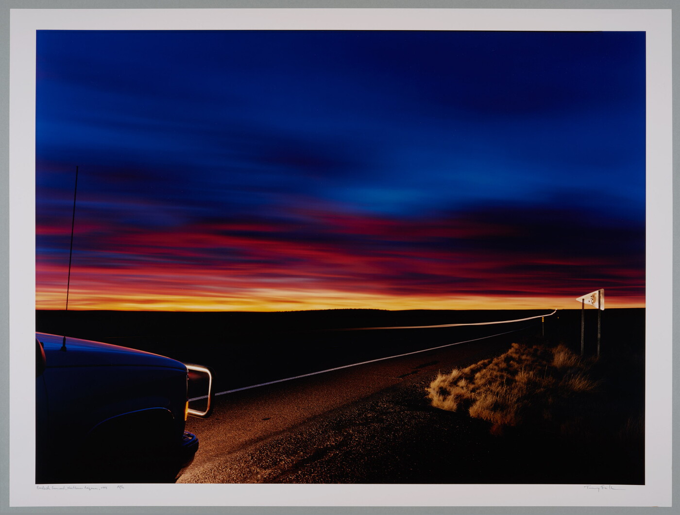 A color photograph of a deep blue sky over a line of orange and yellow sunset in a flat landscape; the lights from the front of a truck in the left corner illuminate part of the road.
