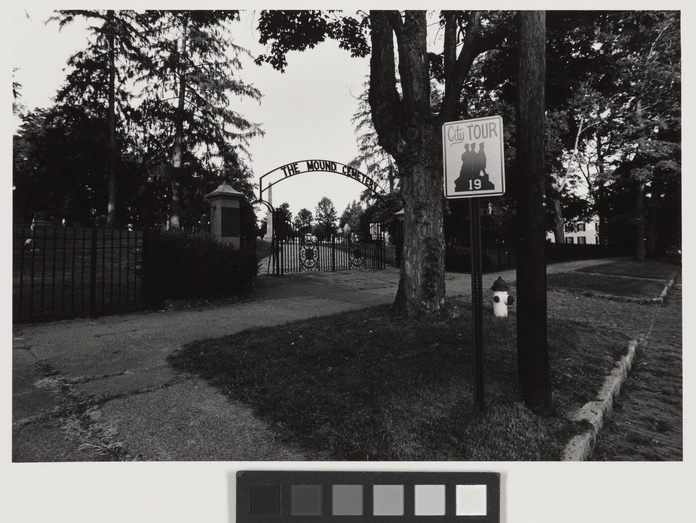 A black-and-white photograph taken from the street of a wrought iron gate a the entrance to a cemetery.