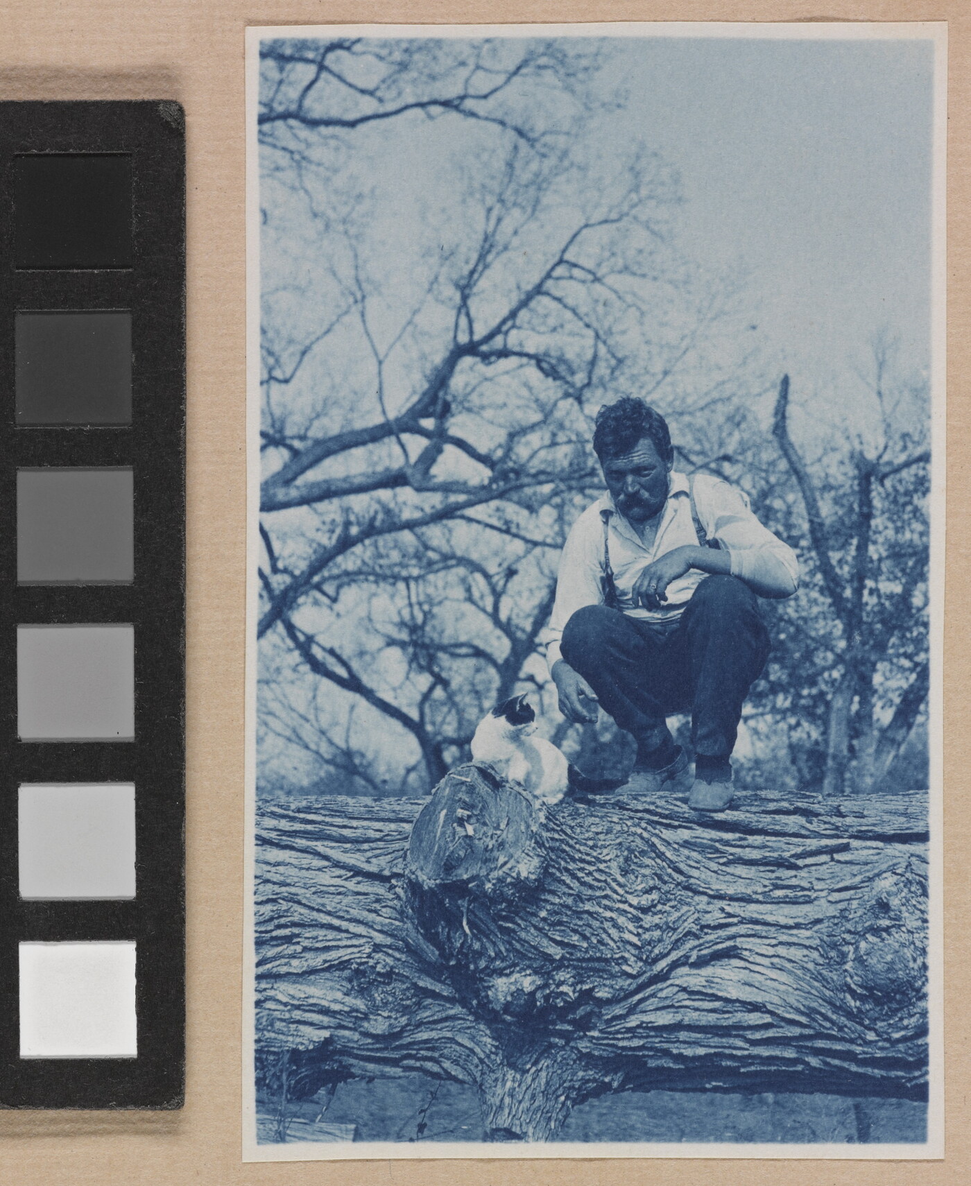 A black-and-white photograph of a White man squatting on a large log next to a white-and-black cat.
