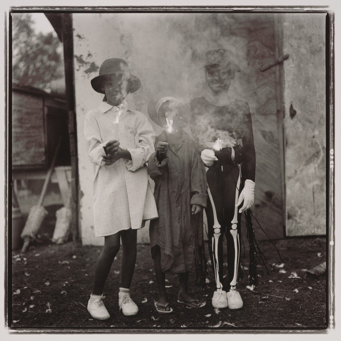 A black-and-white photograph of three Black children: one wearing a hat with a long white shirt, one a hat with a long dark shirt, and one with a hat wearing a skeleton costume; all are holding sparklers with the smoke obscuring their faces.