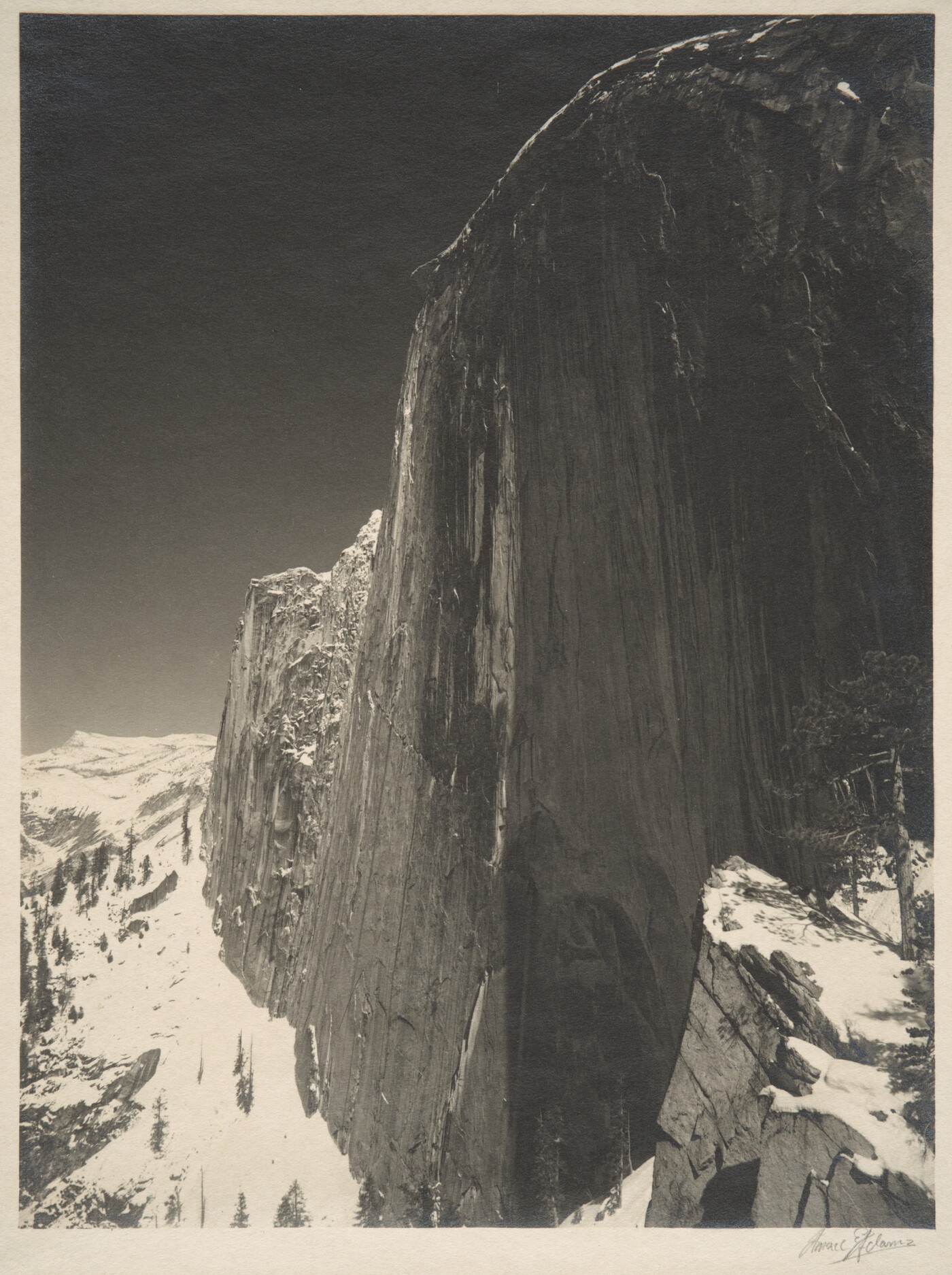 A black-and-white photograph of a steep cliff face and the snow-covered mountains around it.