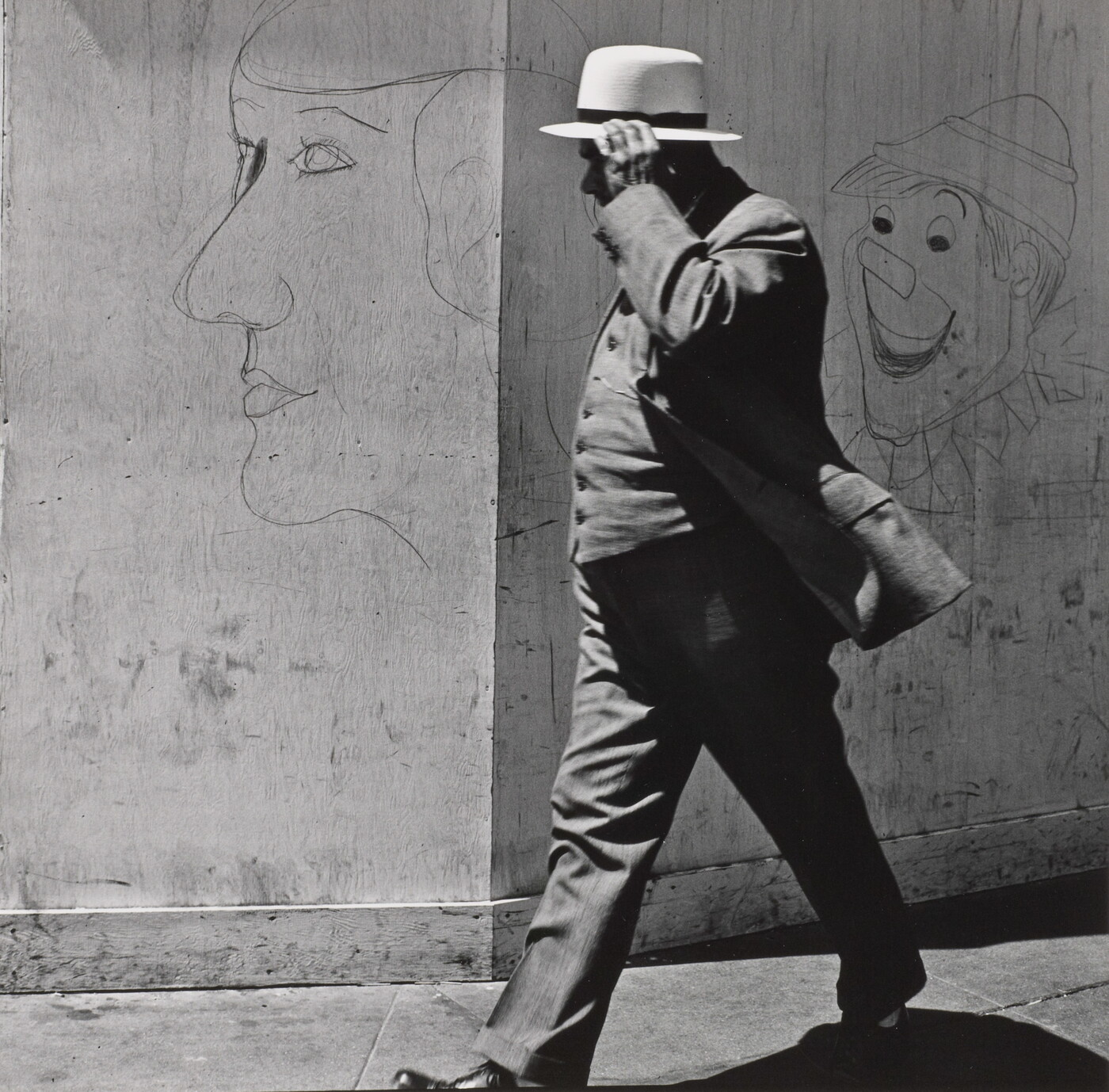 A black-and-white photograph of a man in a three-piece suit and hat walking past a wall covered with graffiti.