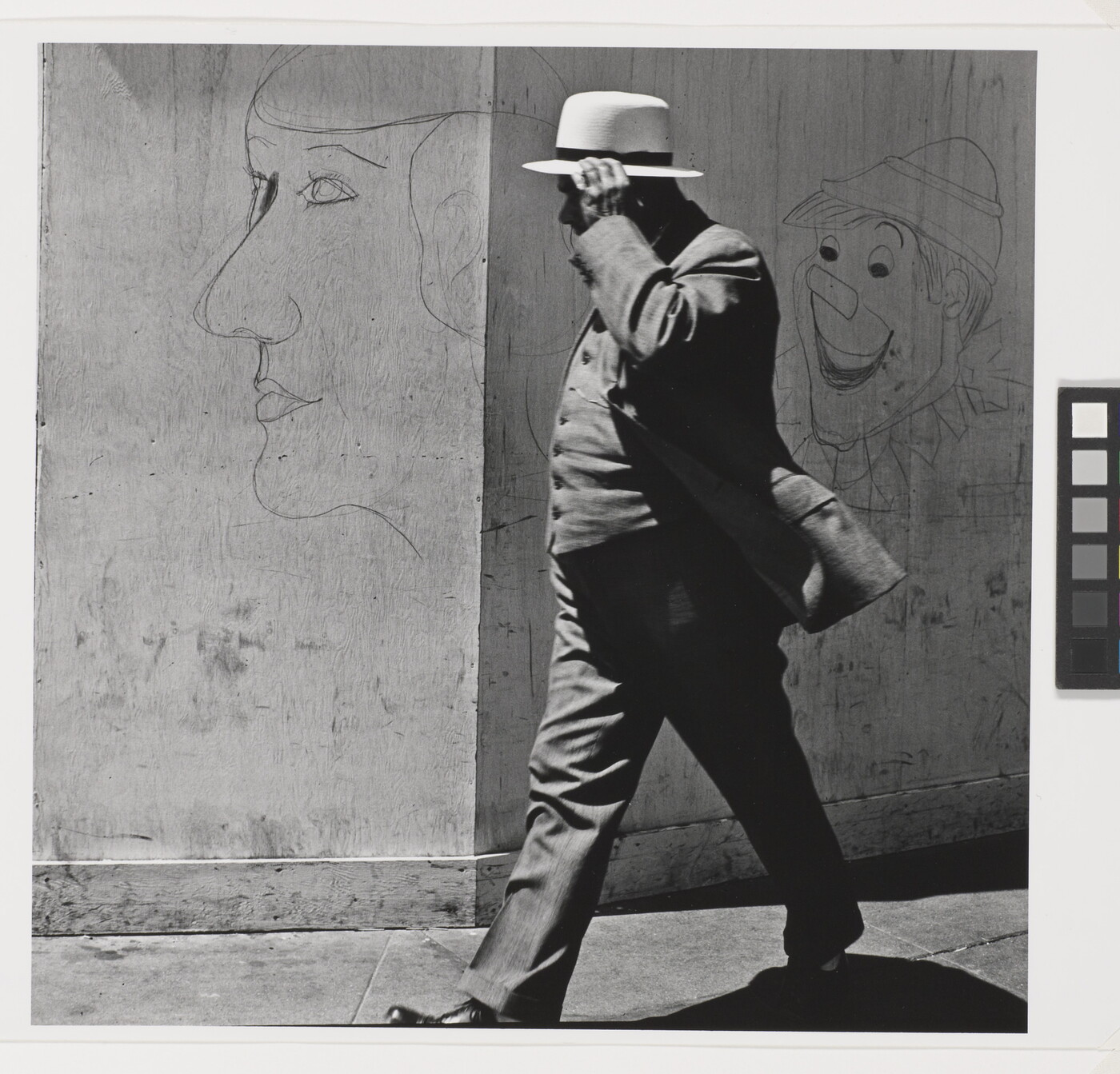 A black-and-white photograph of a man in a three-piece suit and hat walking past a wall covered with graffiti.