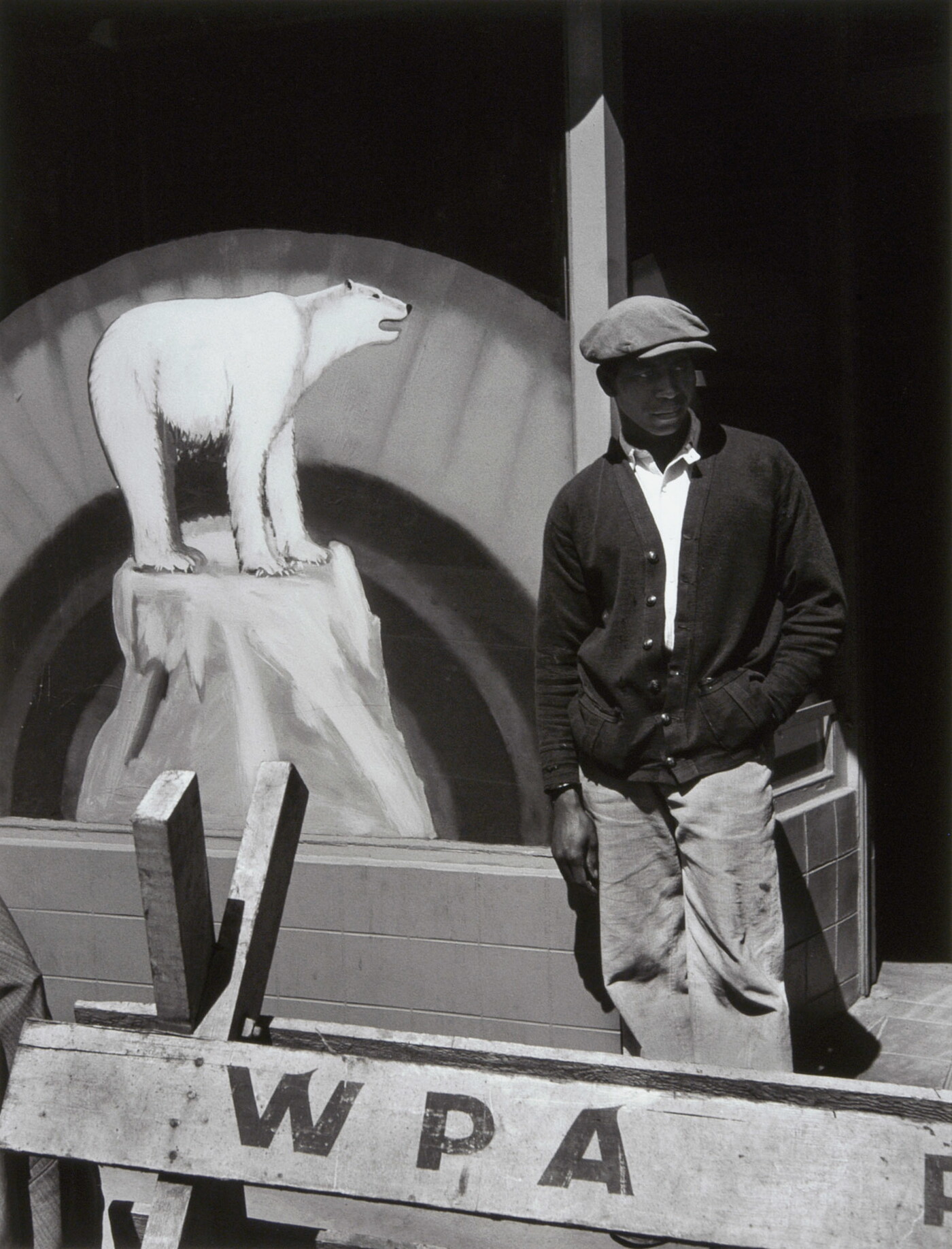A black-and-white photograph of a Black man in a flat cap and dark cardigan behind a wooden barricade that says &quot;WPA&quot; and in front of a sign with a painting of a polar bear.