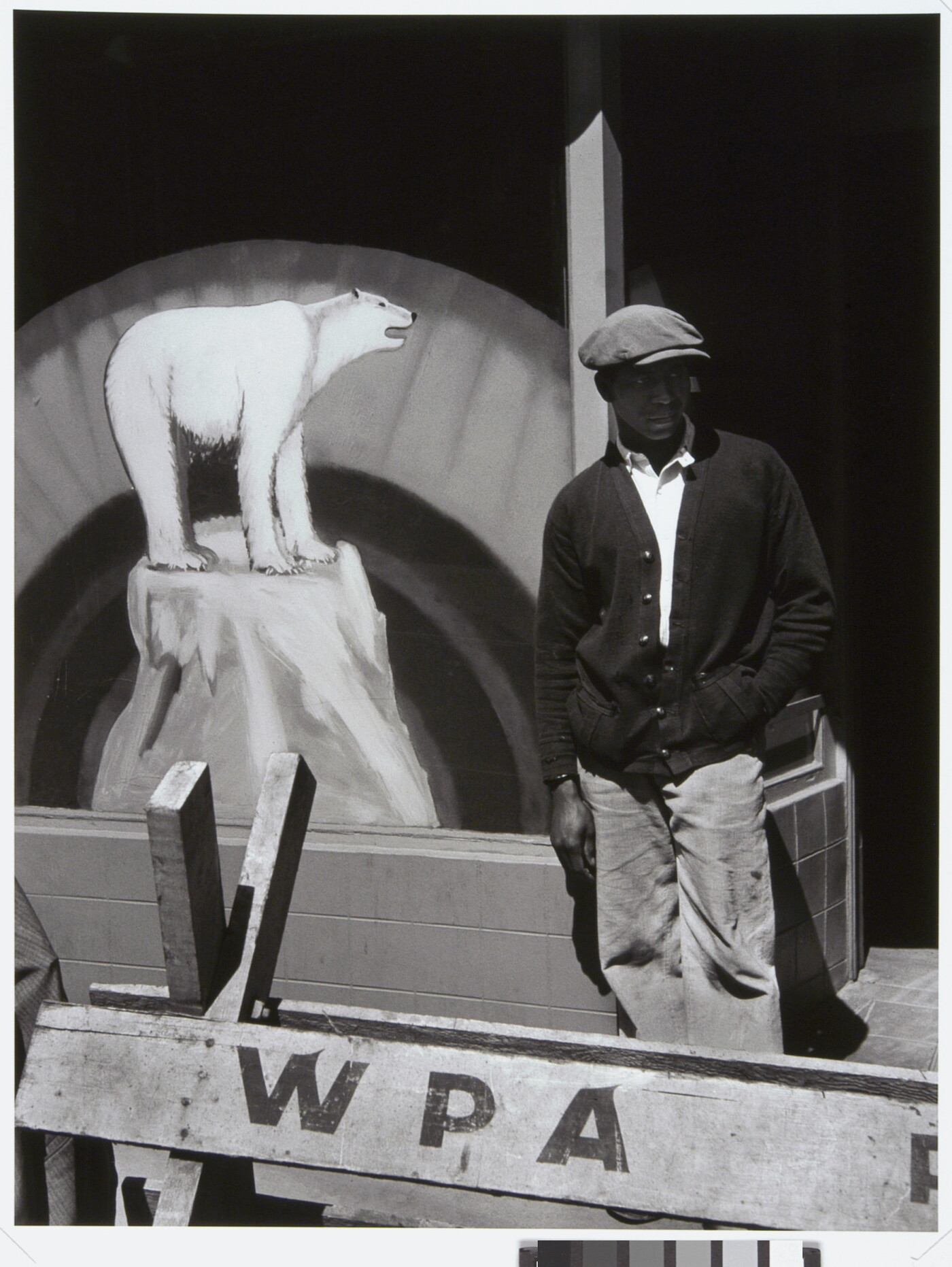 A black-and-white photograph of a Black man in a flat cap and dark cardigan behind a wooden barricade that says &quot;WPA&quot; and in front of a sign with a painting polar bear.