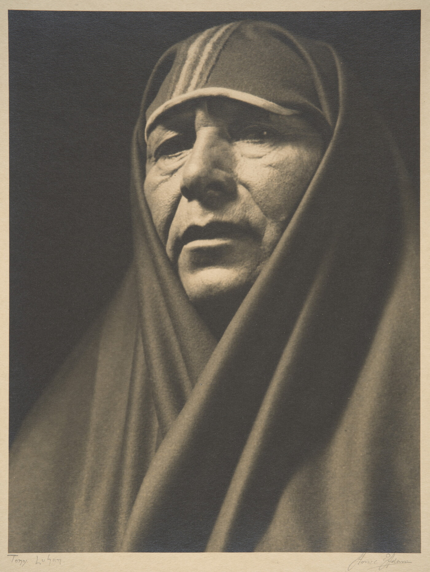 A sepia-toned portrait photograph of an adult Indigenous person with a shawl covering their head and shoulders.