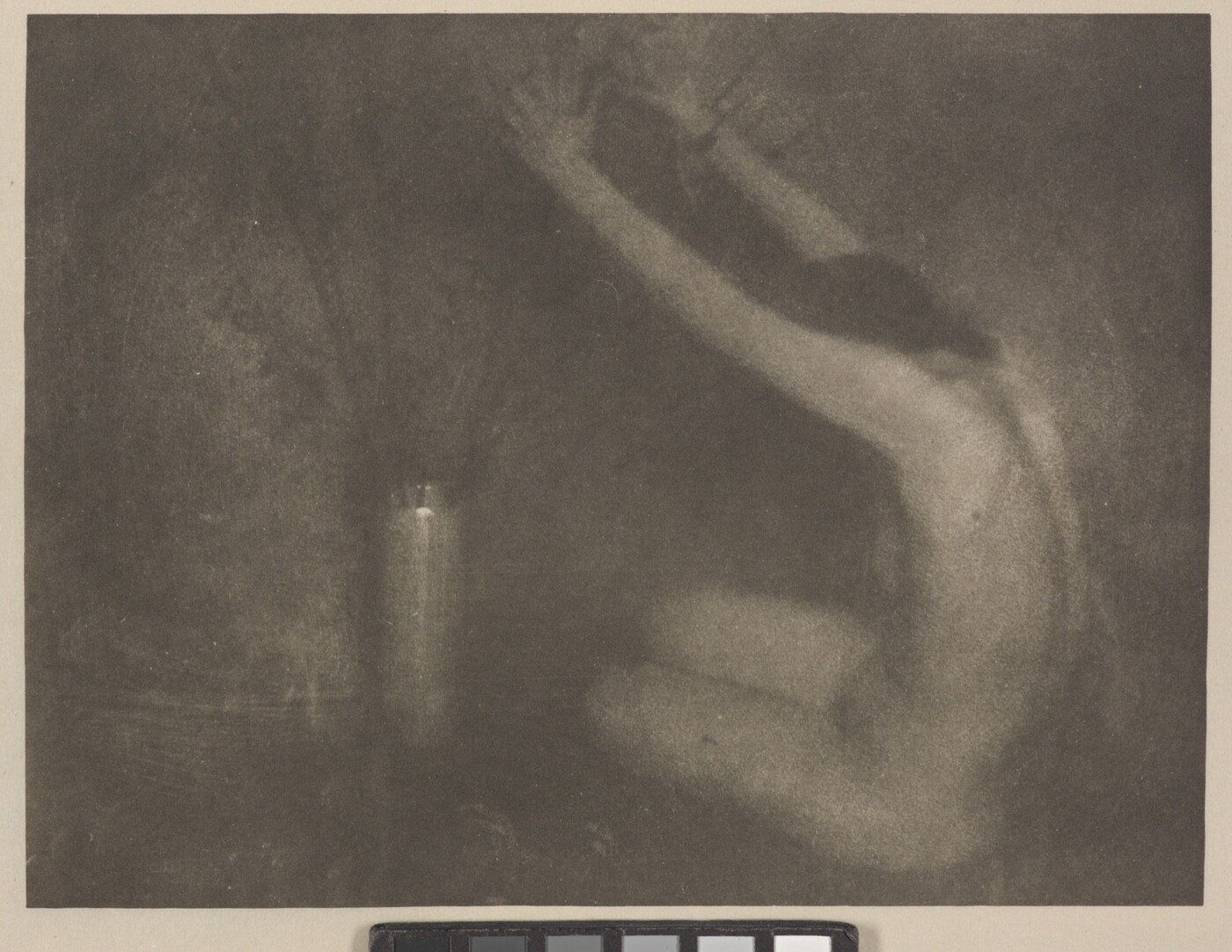 A soft-focus, black-and-white photograph of a nude White woman sitting on the floor next to a vase, her arms raised toward the flowers.