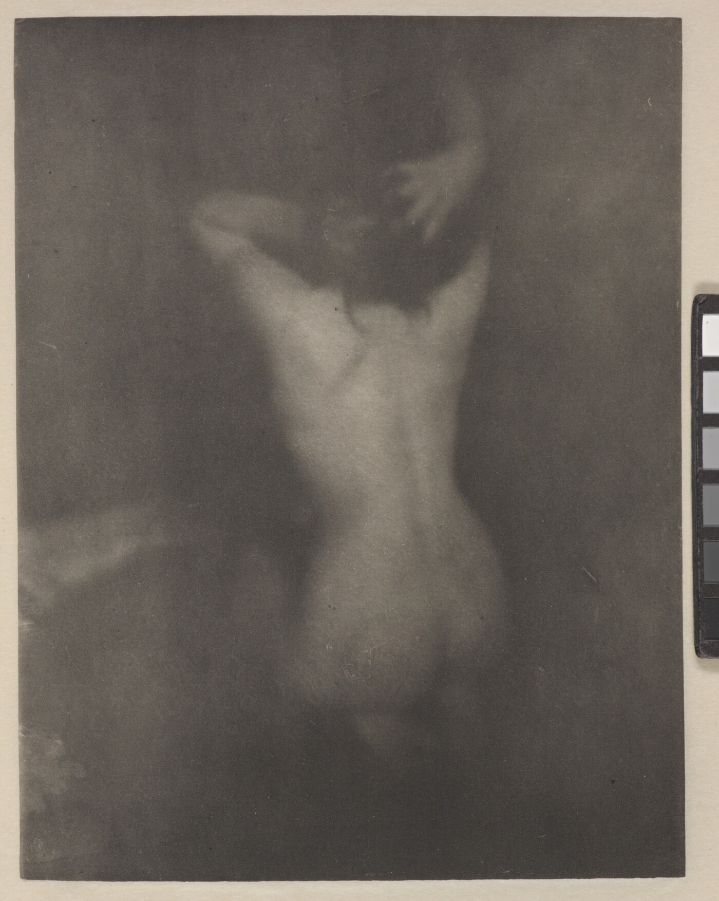 A dark, soft-focus, black-and-white photograph of a nude White woman kneeling with her back to the viewer and her hands in her hair.