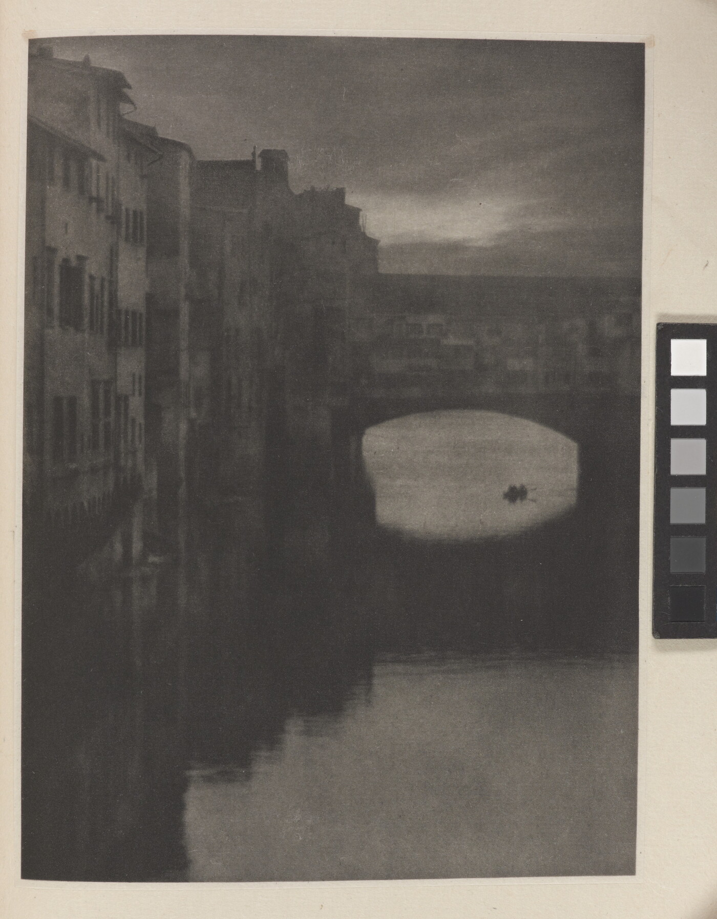 A sepia-toned photograph of a wall of tall buildings lining a river and an enclosed arched bridge stretching over the water.