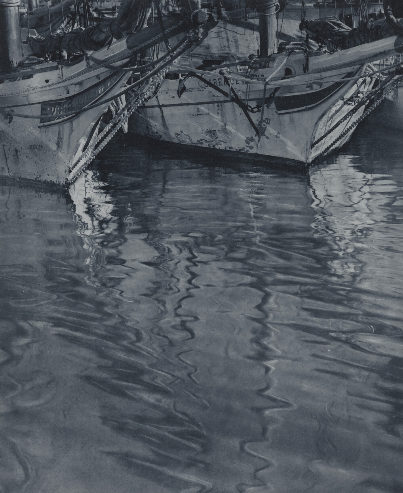 A black-and-white photograph of the ends of two docked sailboats floating on rippling water.