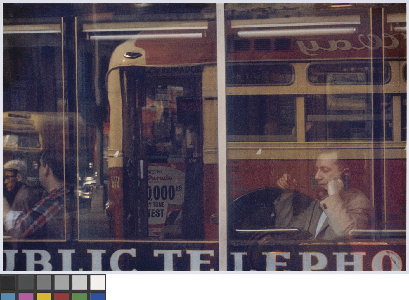 A color photograph of a White man sitting at a cafe window speaking on a phone, with reflections of a passing bus in the glass.