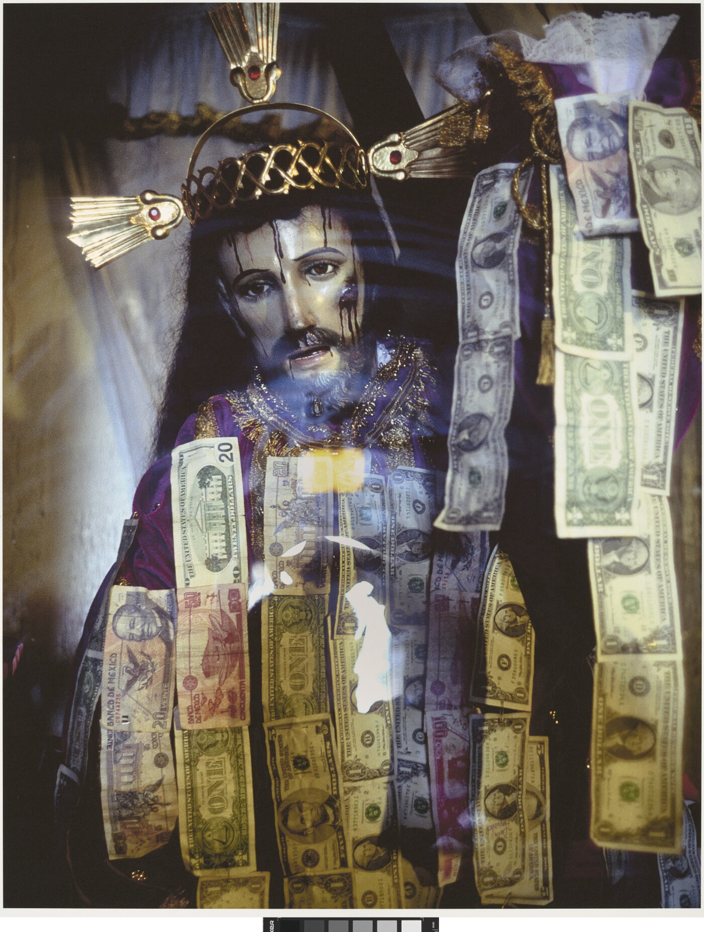 A color photograph of a sculpture of Jesus Christ wearing a crown of thorns behind glass that has strips of dollar bills hanging on it.