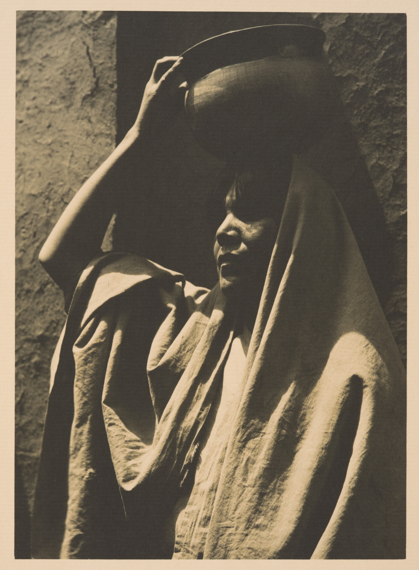 A black-and-white photograph of an Indigenous woman, face mostly in shadow, head and shoulders covered in a blanket or shawl, using her hand to balance a pot on her head.