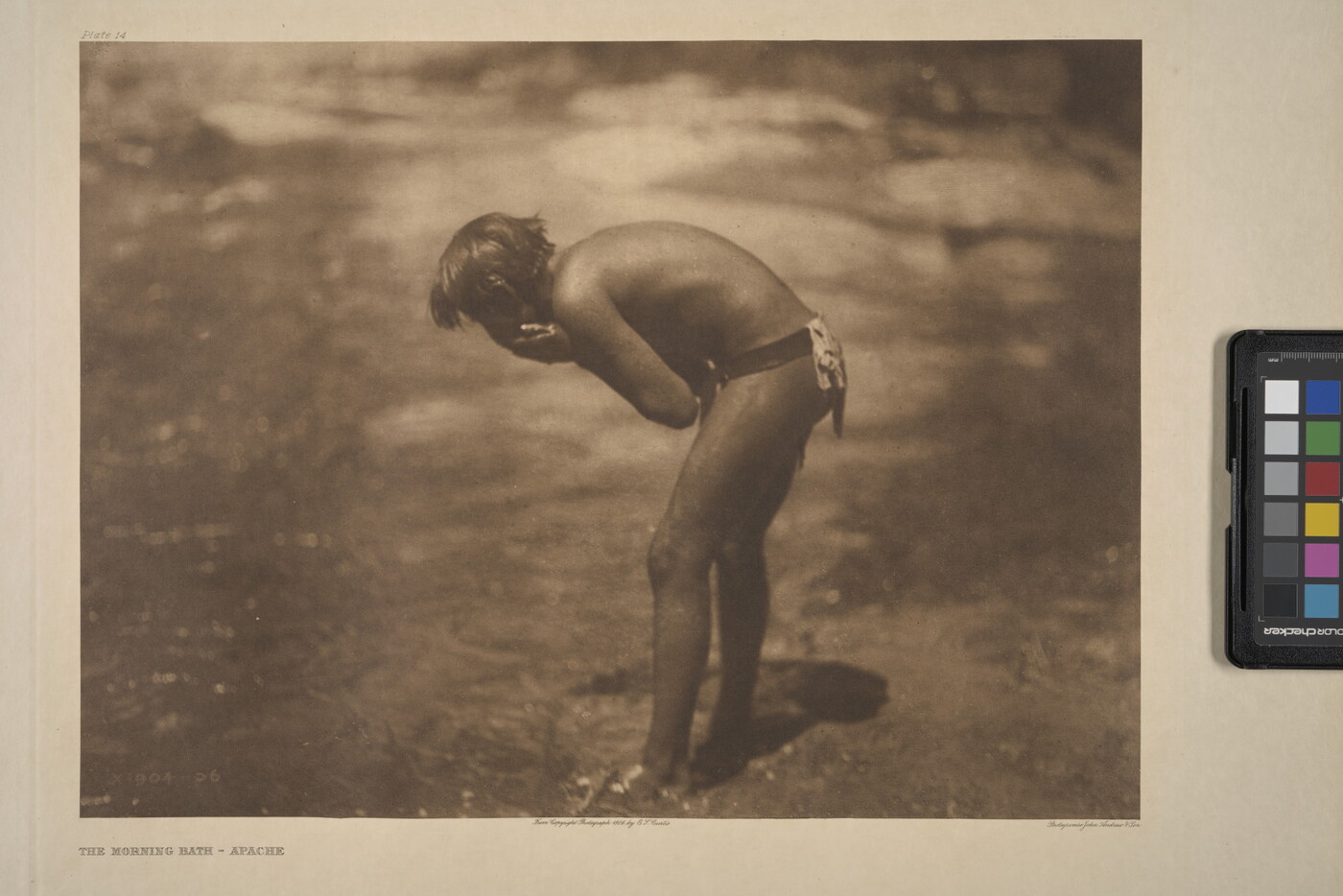 A sepia-toned photograph of a young Indigenous person leaning over a riverbank washing their face.
