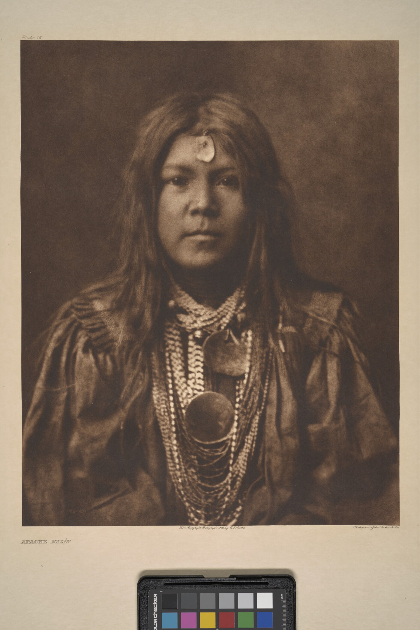 A sepia-toned portrait photograph of a young Indigenous person with long hair, a piece of jewelry on their forehead, multiple stings of beads around their neck, and wearing cultural dress.