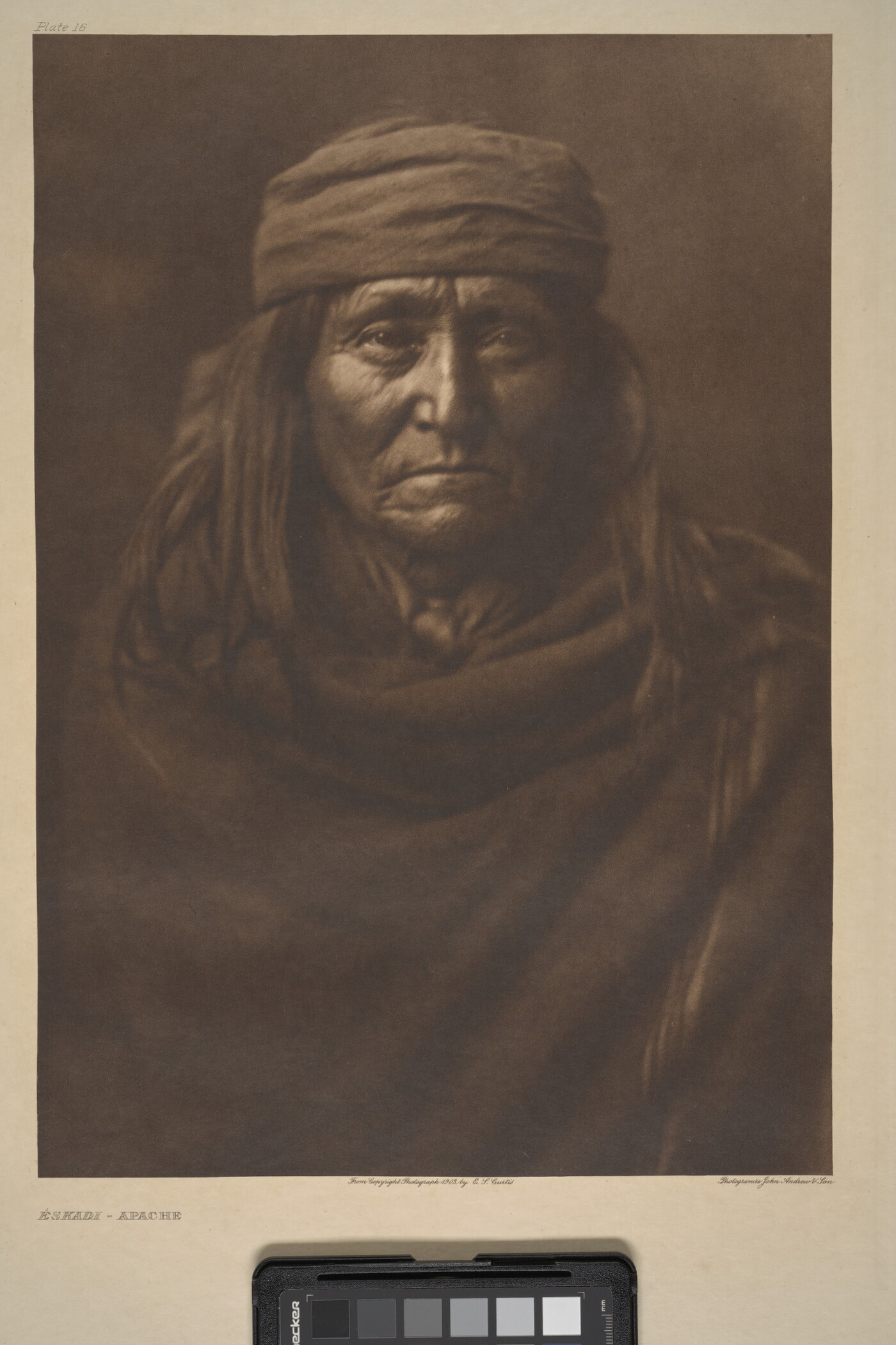 A sepia-toned portrait photograph of an older Indigenous person with long hair, wearing a cap, and wrapped in a solid-colored cloth.