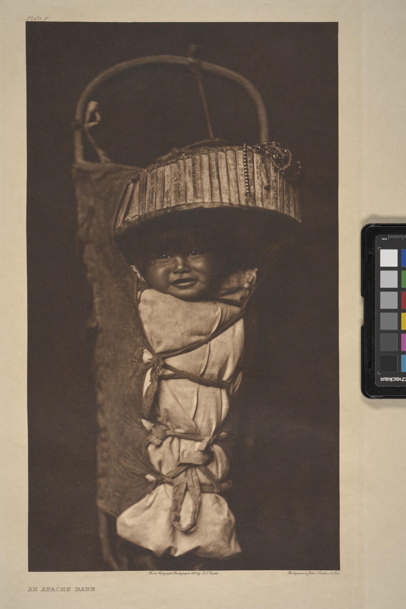 A sepia-toned photograph of a smiling Indigenous baby who is strapped into a traditional cradleboard.