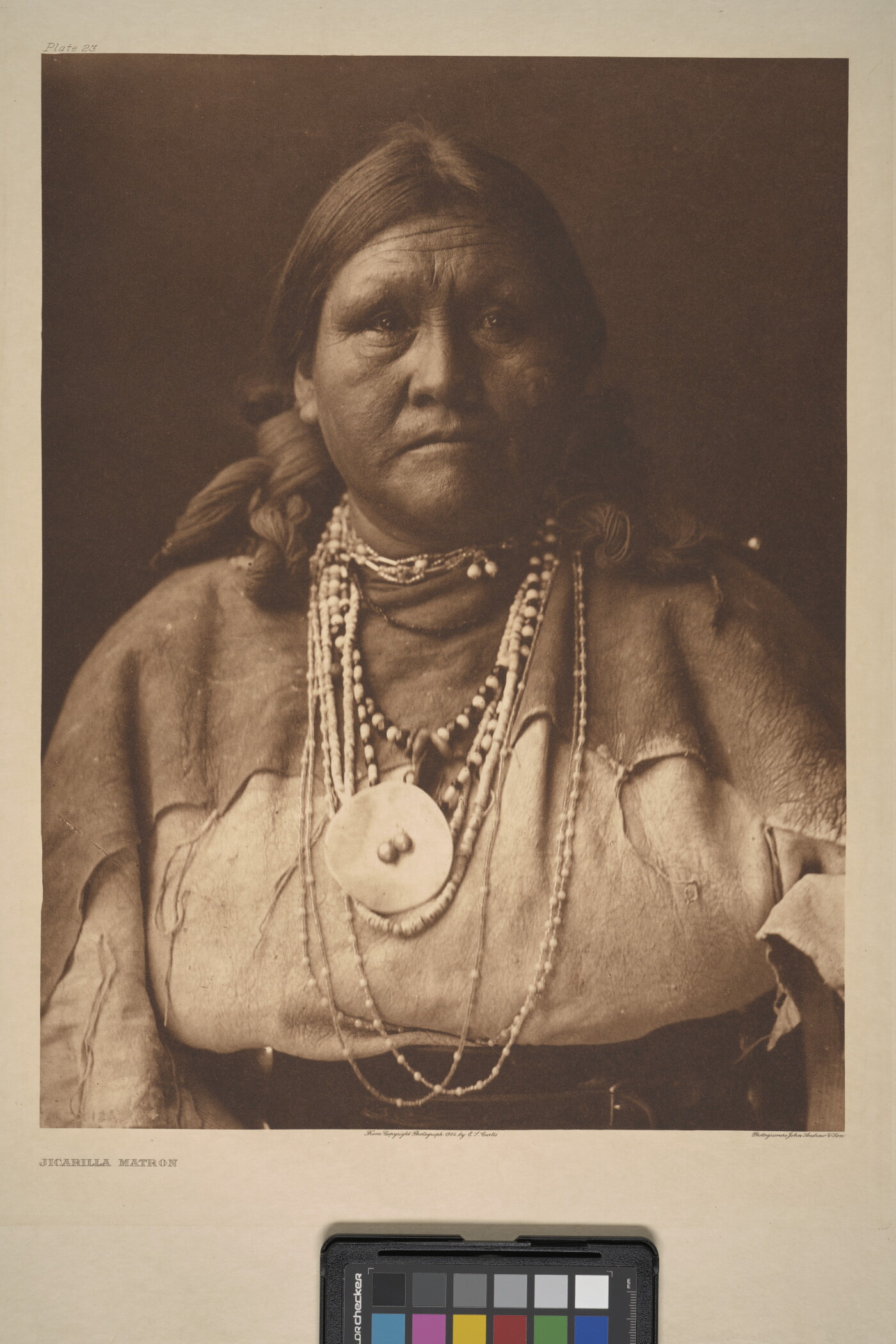 A sepia-toned portrait photograph of an older Indigenous woman with her hair bound on both sides of her head and wearing several beaded necklaces and traditional clothing.