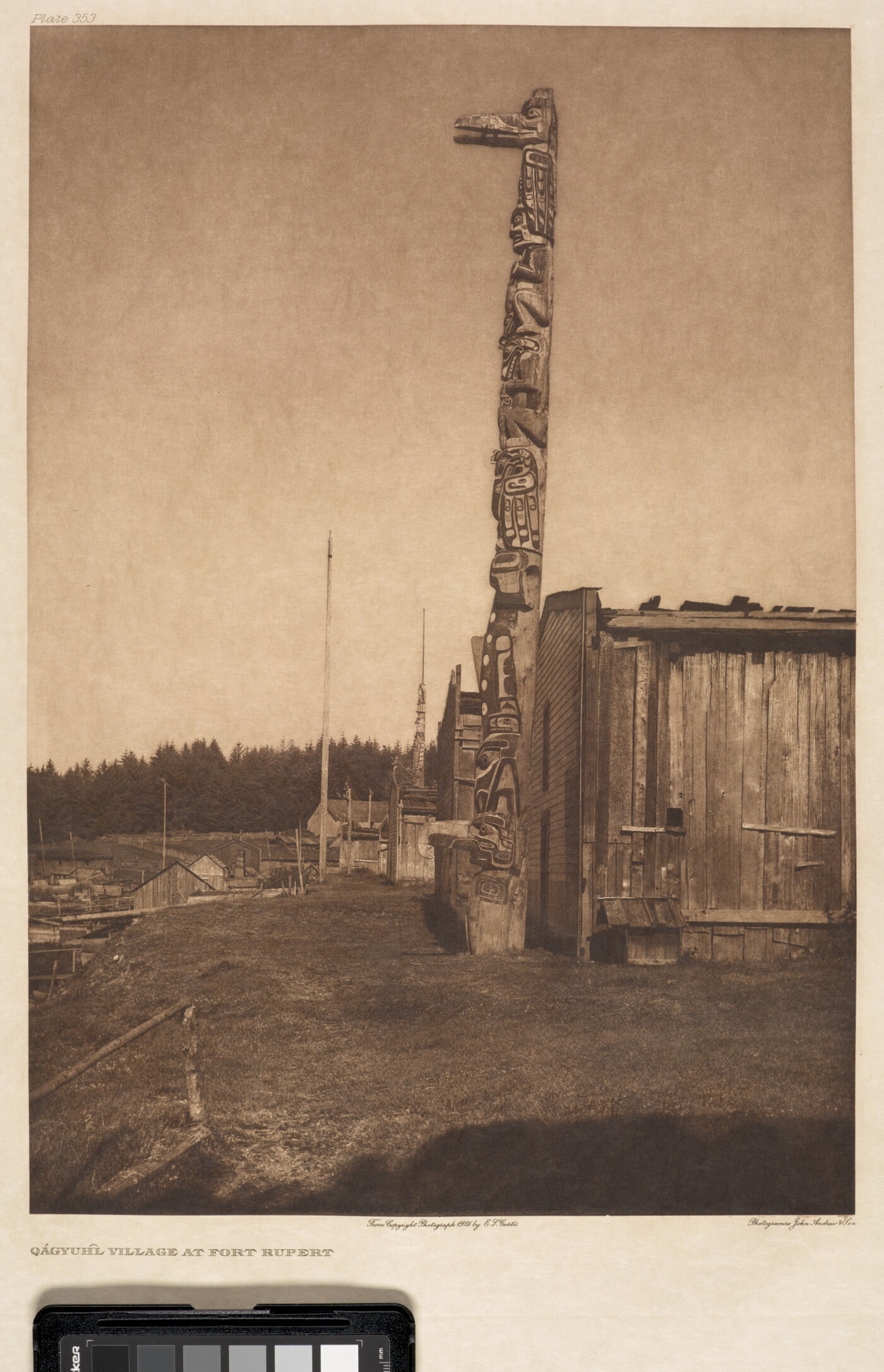 A sepia-toned photograph of a tall totem pole next to a wooden building with a forest in the background.