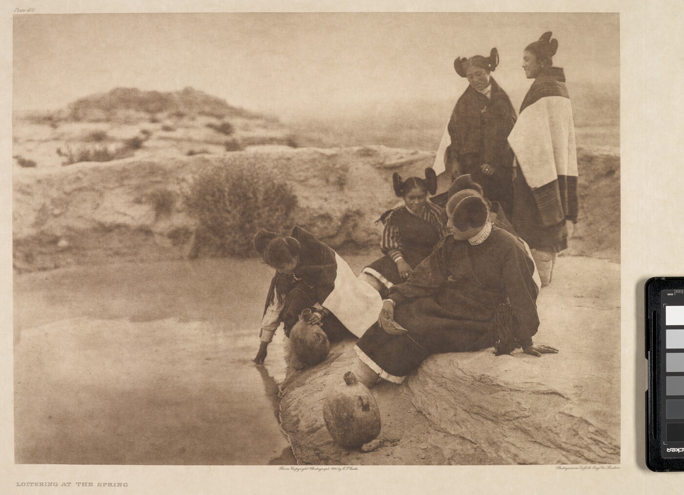 A sepia-toned photograph of a group of five Indigenous women wearing traditional dress sitting and standing on the edge of a spring.