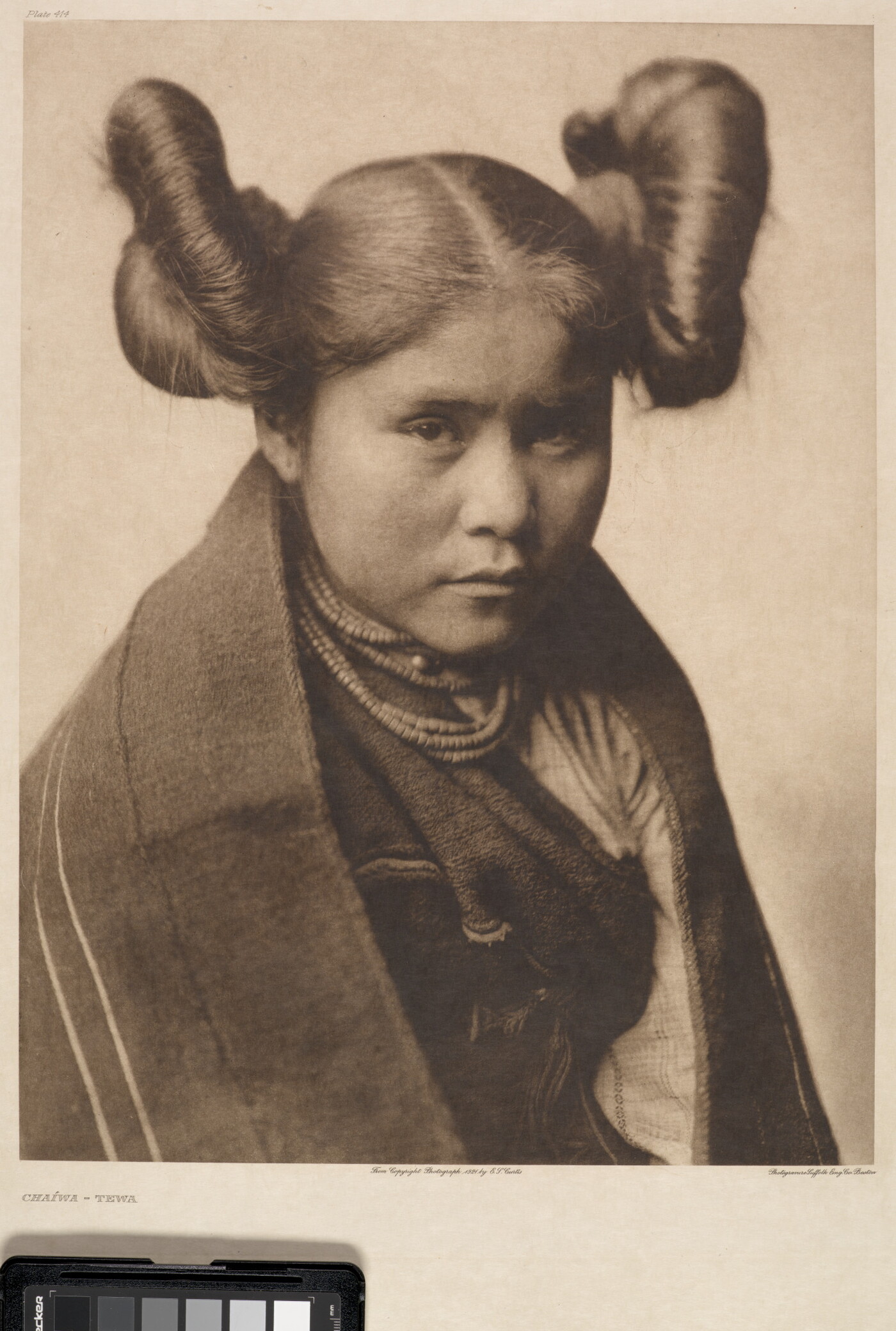 A sepia-toned portrait photograph of a young Indigenous woman with her hair split into two large buns on both sides of her head wearing traditional clothes and necklaces.