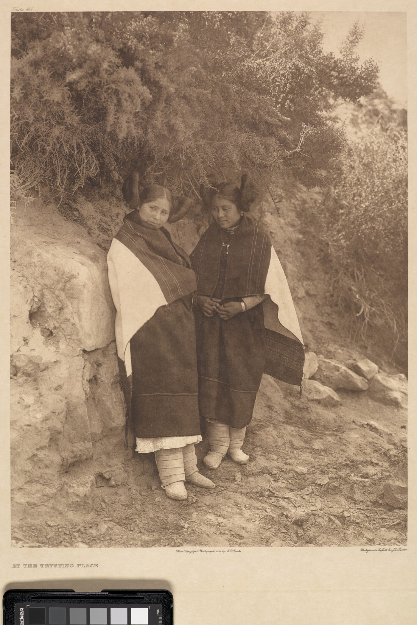 A sepia-toned photograph of two Indigenous women in traditional clothing leaning on a rock that has vegetation growing off the top of it.