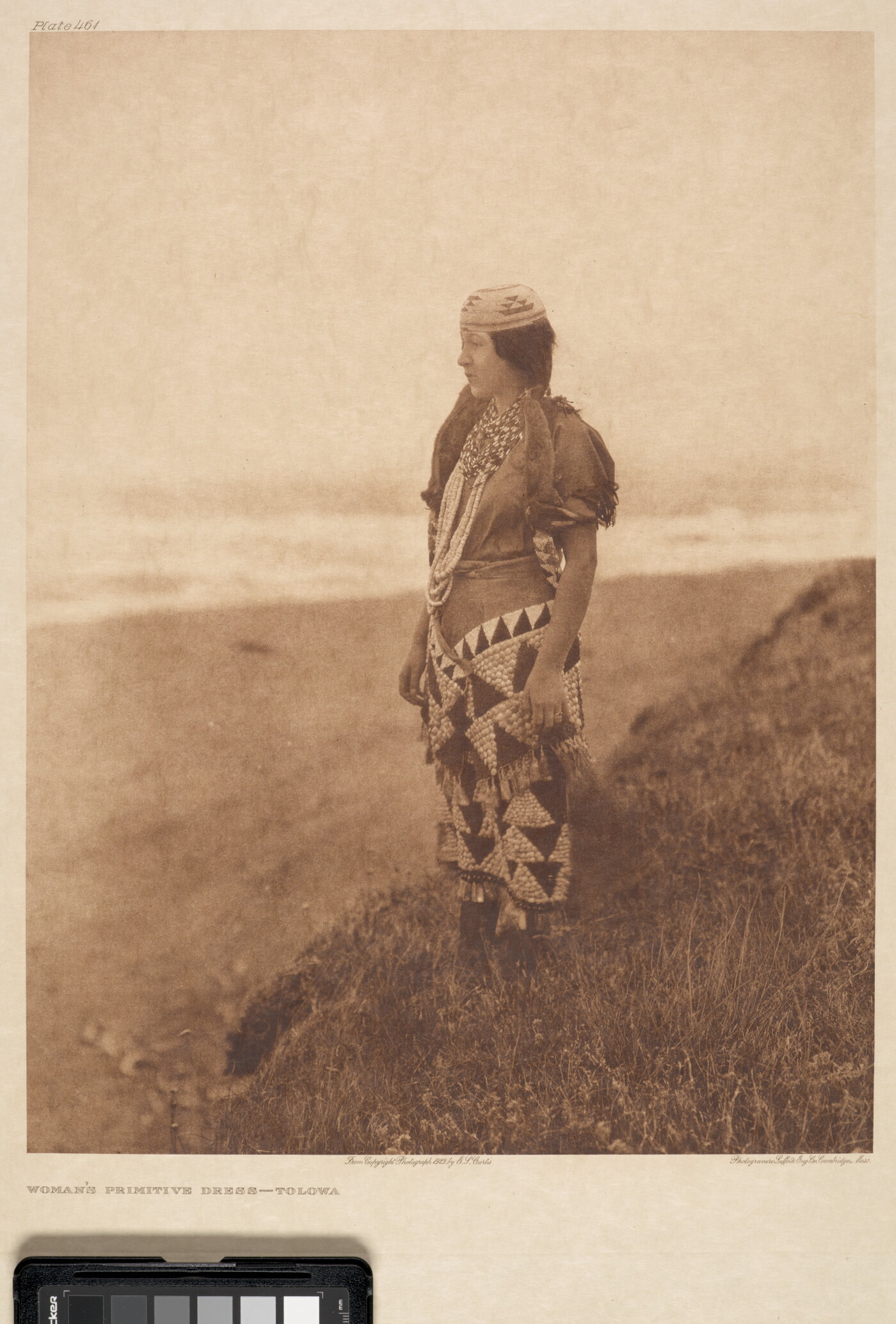 A sepia-toned photograph of a young Indigenous woman wearing traditional dress standing on a hill overlooking a plain.
