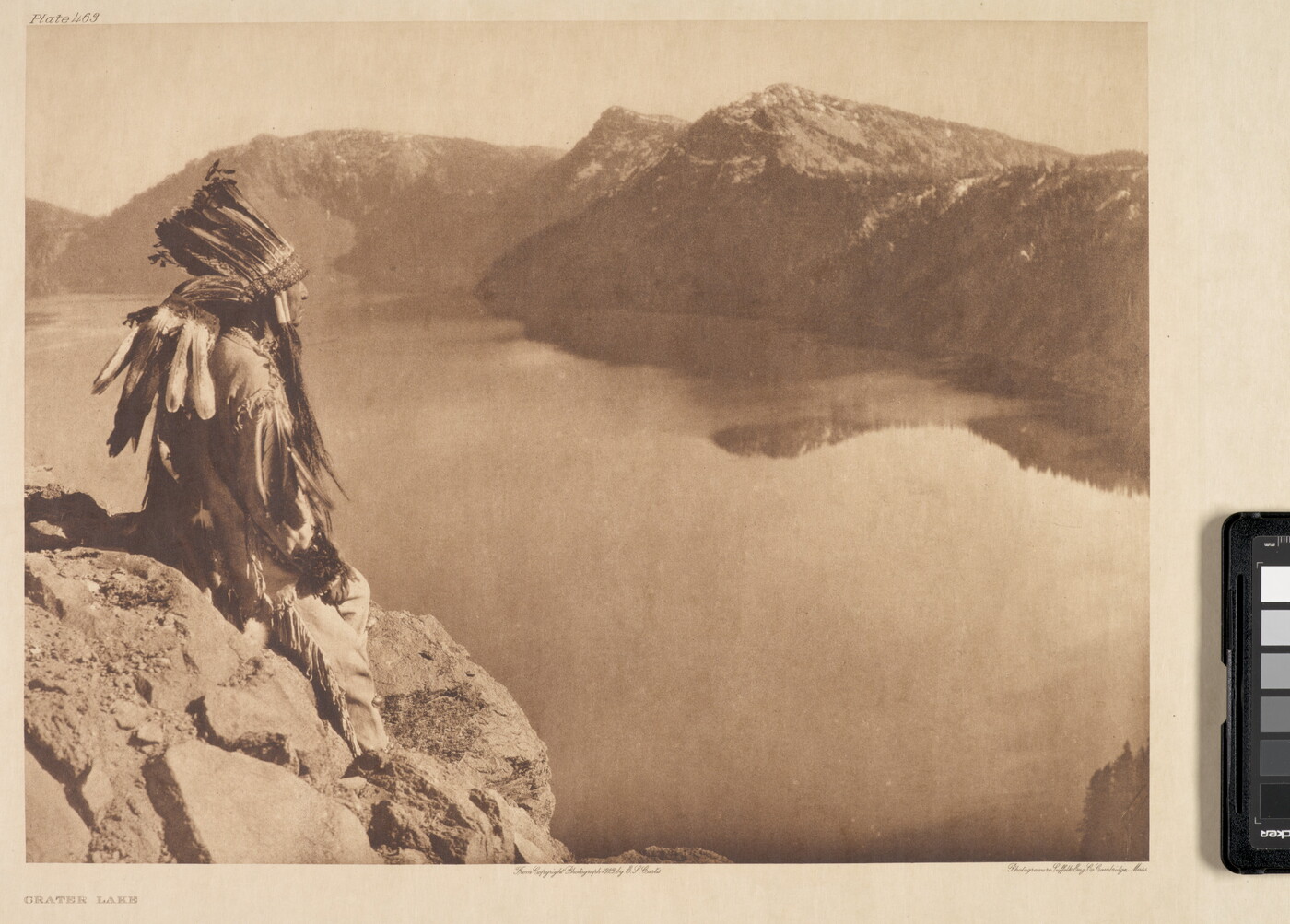 A sepia-toned photograph of an Indigenous man in traditional clothing and a feathered headdress sitting on a rock high above a lake at the base of mountains.