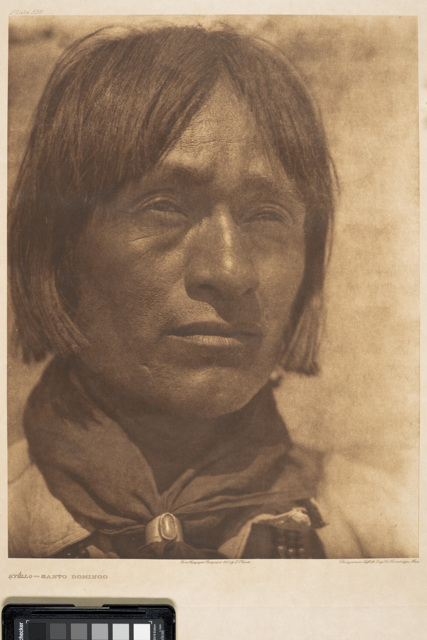 A close-up, sepia-toned portrait photograph of an Indigenous man with hair to his jawline, wearing a scarf around his neck held by a silver concho-style scarf slide.