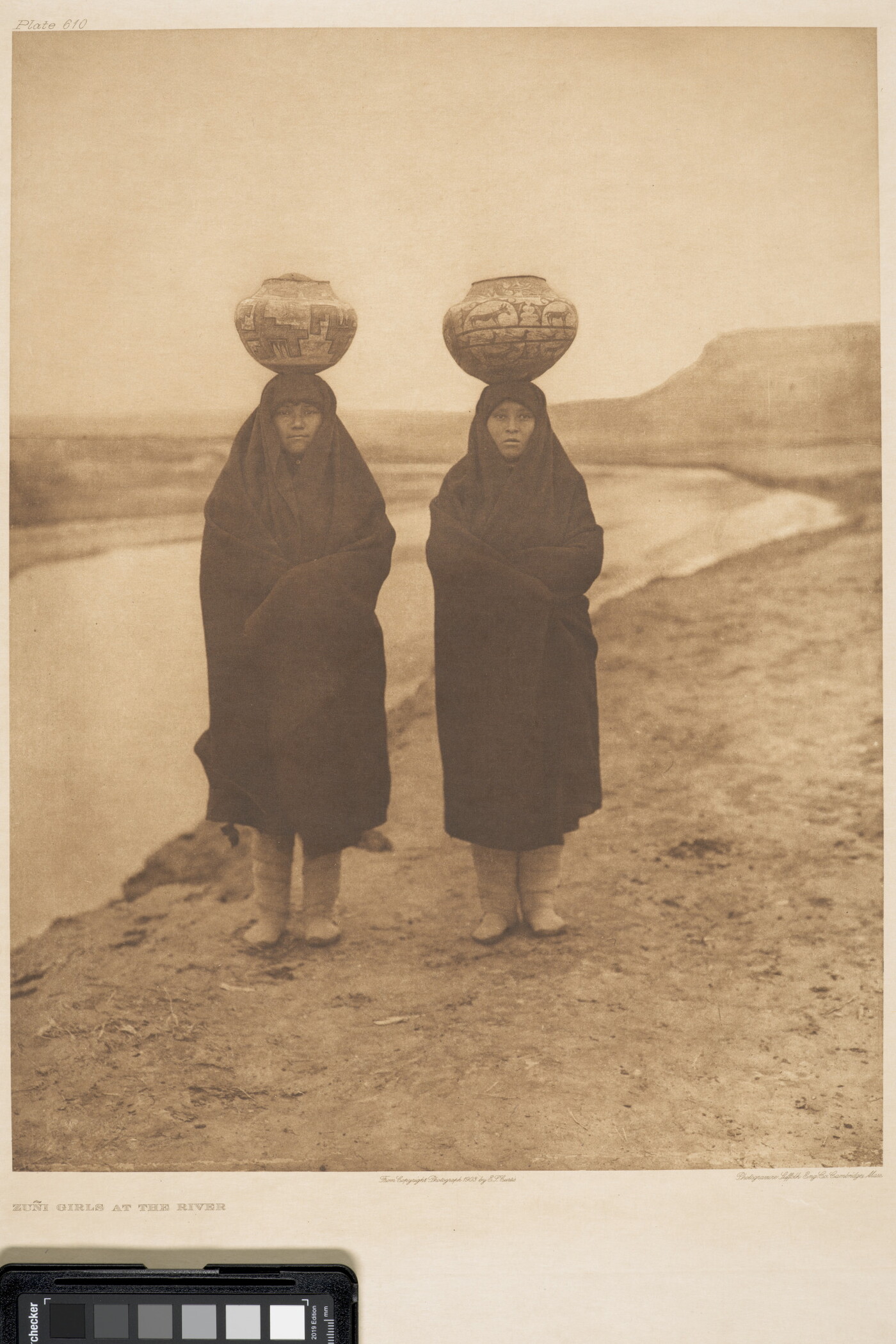 A sepia-toned photograph of two Indigenous women wrapped in blankets standing on a riverbed with water jugs balanced on their heads.