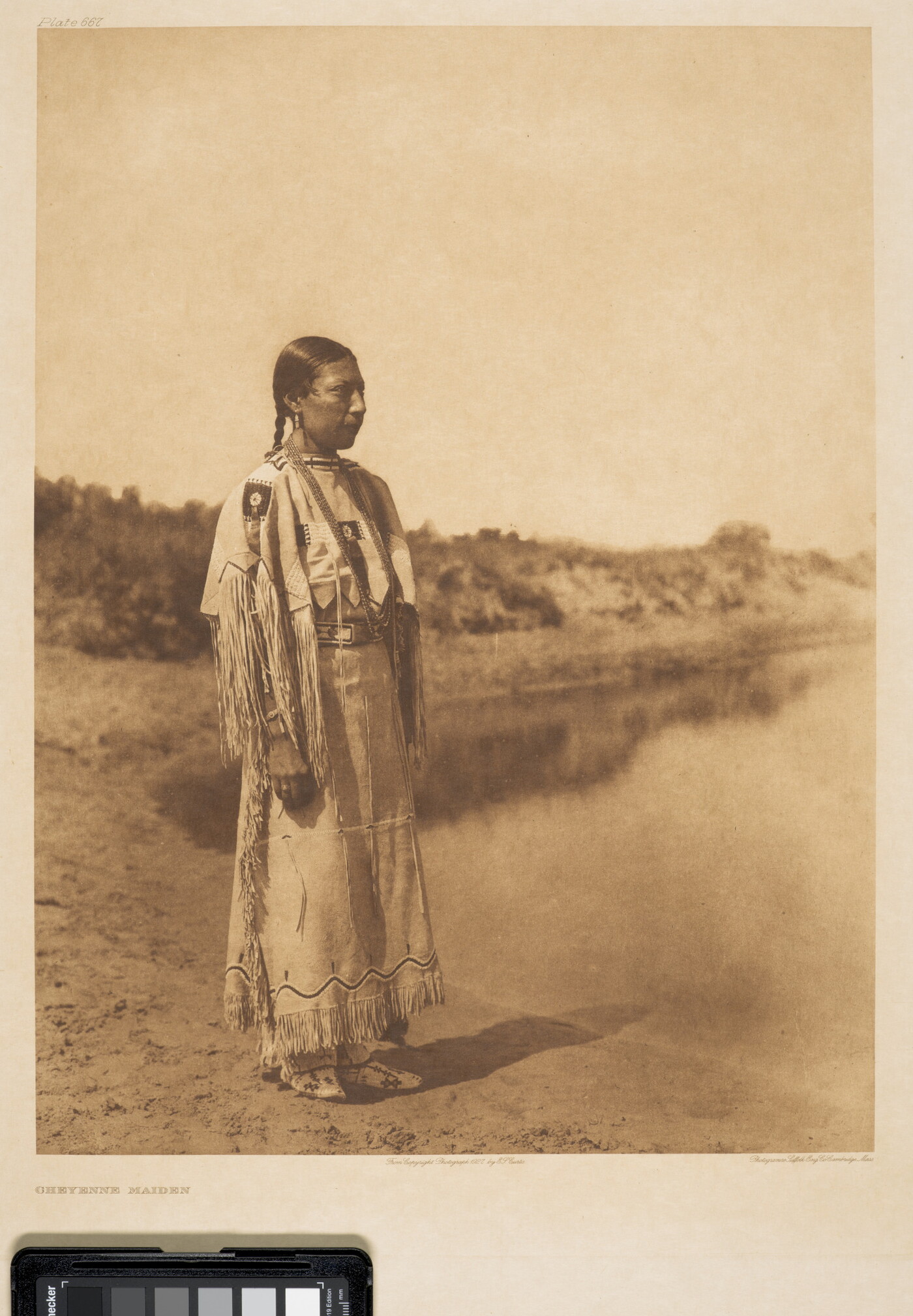 A sepia-toned photograph of an Indigenous woman with her hair pulled back into a braid, wearing fringed clothing that looks to be made of animal skins, standing on a shoreline.