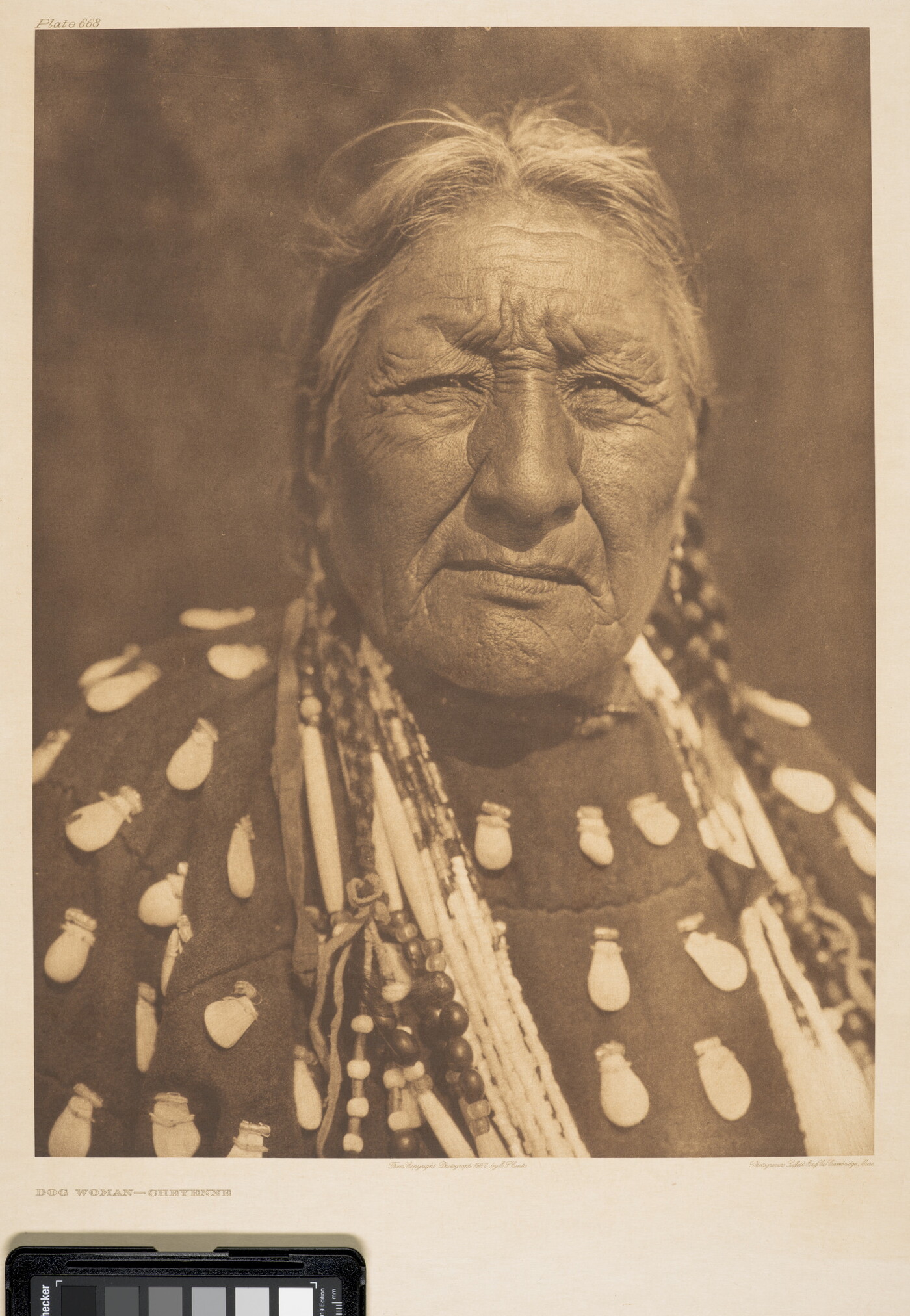 A sepia-toned portrait photograph of an older Indigenous woman wearing a braids, elk-tooth shirt, and multiple necklaces.