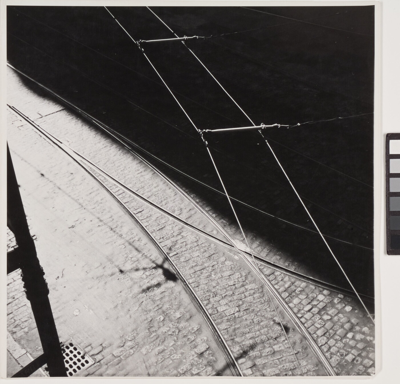 A black-and-white abstract photograph of tram tracks, a brick street, and wires divided diagonally by a hard shadow.