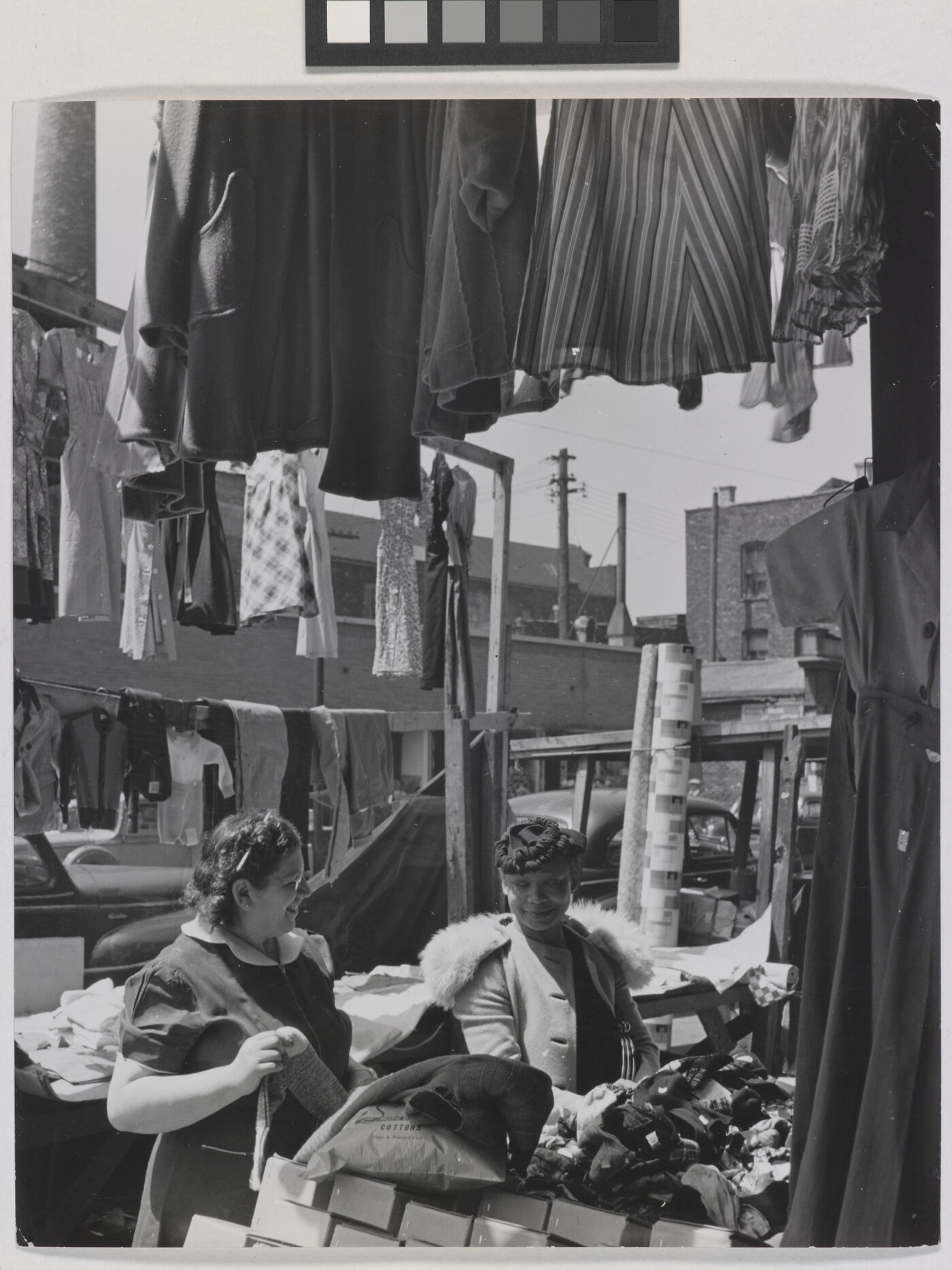 A black-and-white photograph of two light- to medium-skinned women smiling at each other in an open-air market, surrounded by clothing and boxes.