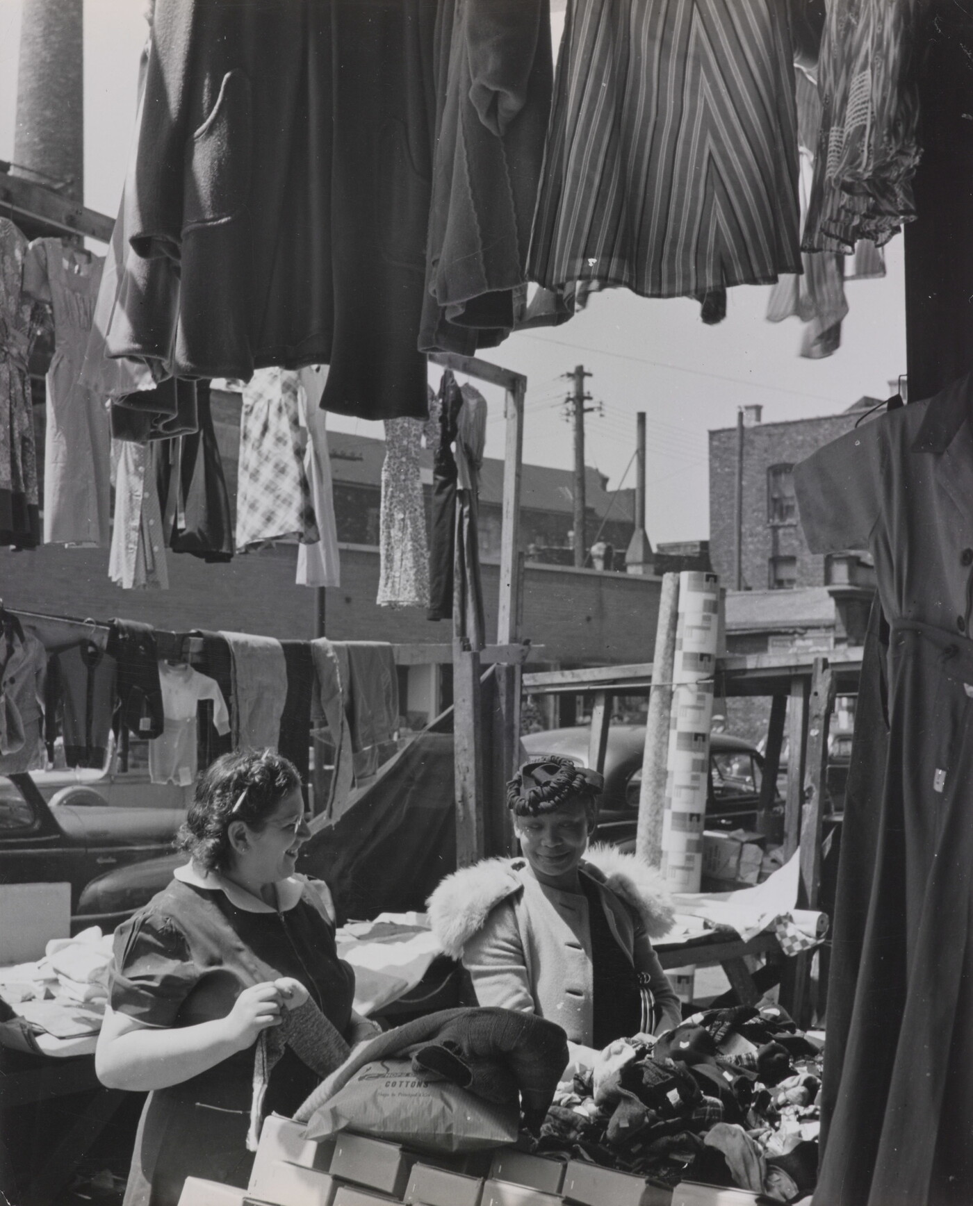 A black-and-white photograph of two light- to medium-skinned women smiling at each other in an open-air market, surrounded by clothing and boxes.