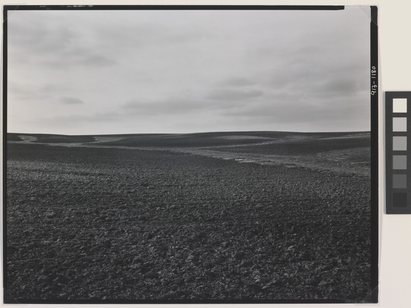 A black-and-white photograph of a dirt landscape with subtle hills under a cloudy sky.