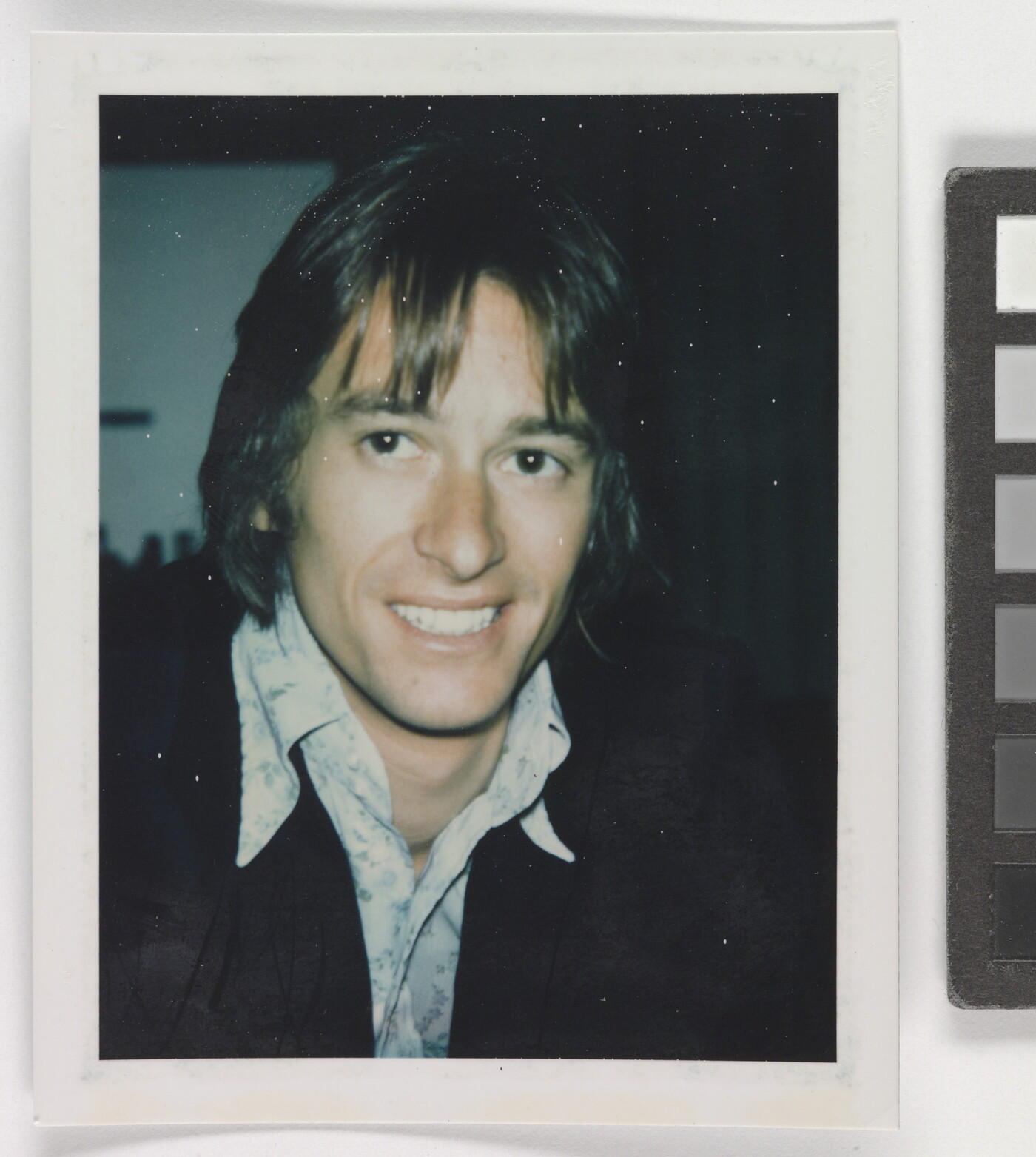 An informal color photograph of a smiling White man from the shoulders up with longish hair, wearing an open collar shirt and dark jacket.